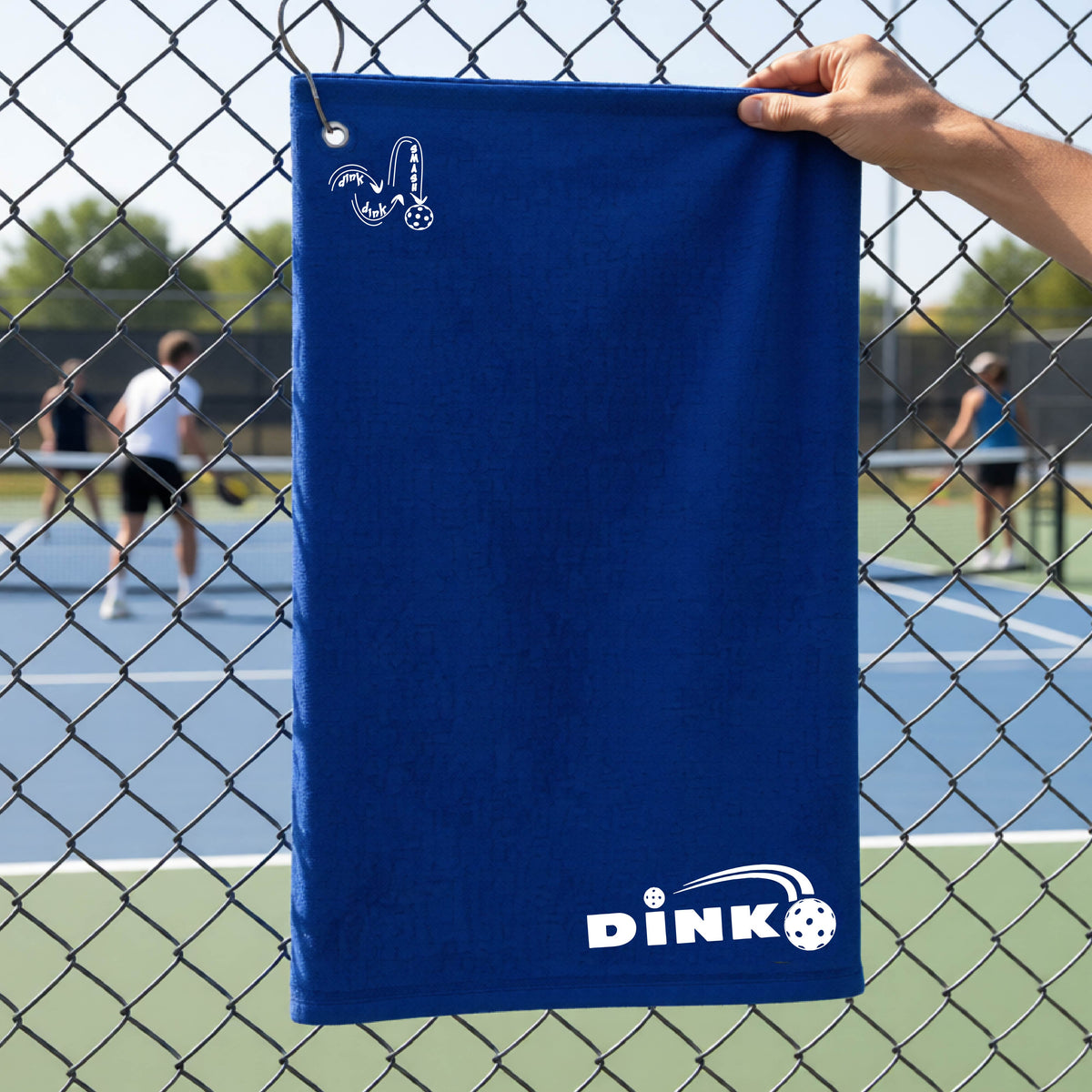 A close up of a royal blue pickleball court towel with a log of 'DINK DINK SMASH' in the upper left corner and a pickleball design of text 'DINK' with a white pickleall in flight, with a pickleball court in the background. 