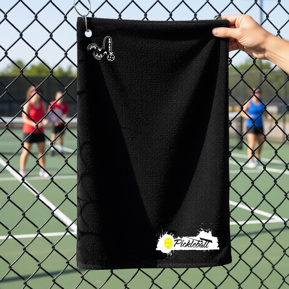 A black pickleball terry cloth towel in front of a chain-link fence at a pickleball court, The towel features a white and black Pickleball splatter graphic near the bottom rigfht and a white DINK DINK SMASH Pickleball icon near the top left.