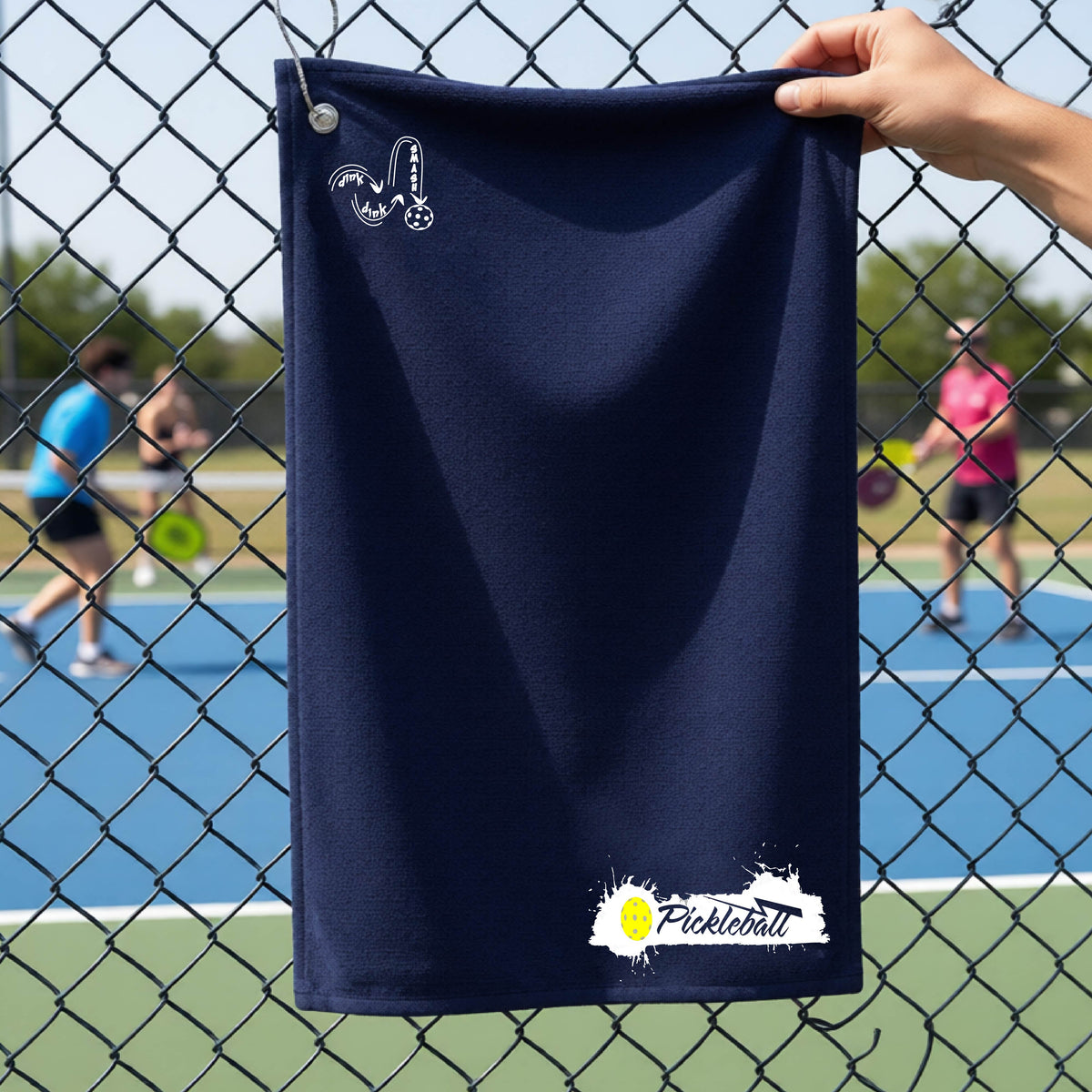 A navy blue pickleball terry cloth towel in front of a chain-link fence at a pickleball court, The towel features a white and black Pickleball splatter graphic near the bottom rigfht and a white DINK DINK SMASH Pickleball icon near the top left.
