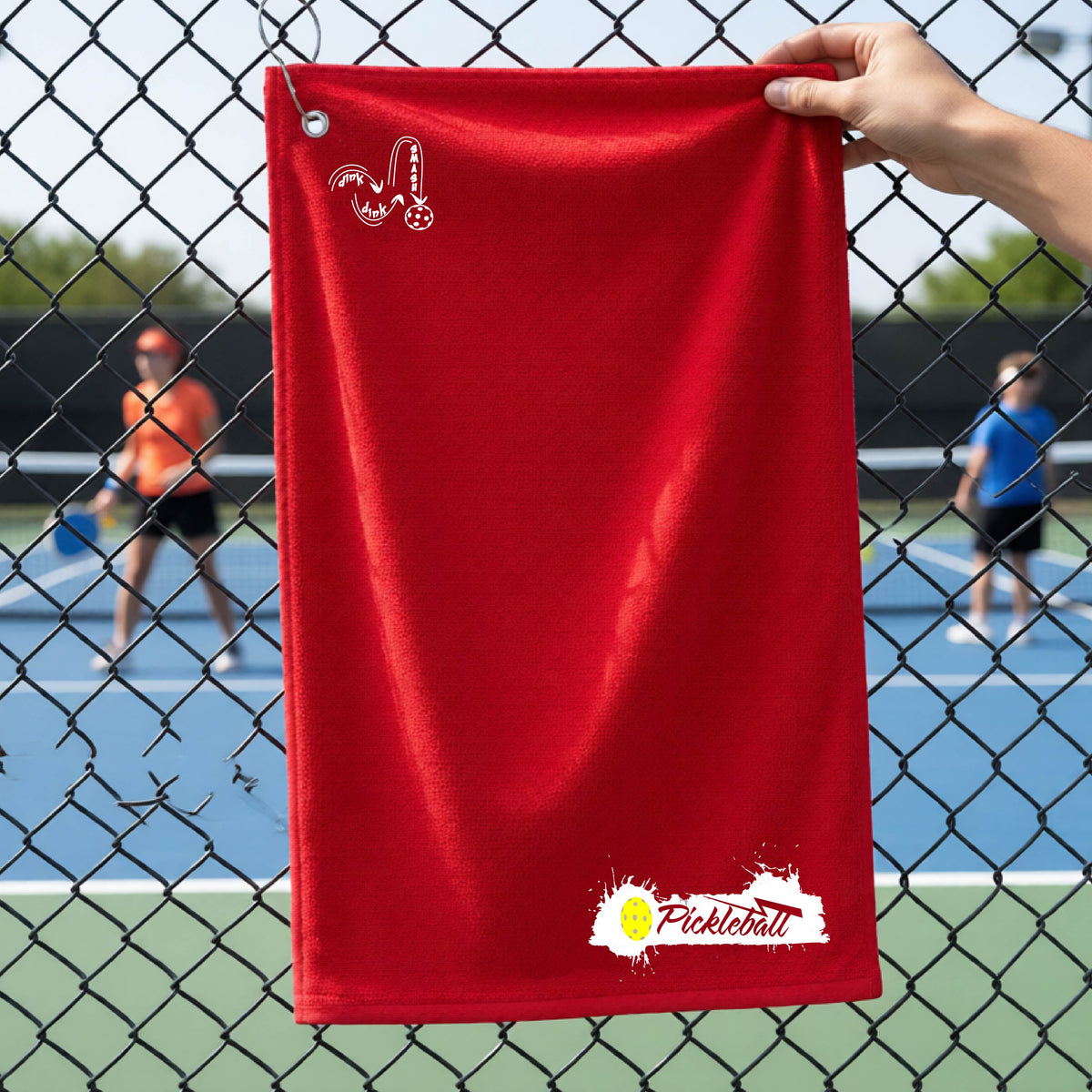 A red pickleball terry cloth towel in front of a chain-link fence at a pickleball court, The towel features a white and black Pickleball splatter graphic near the bottom rigfht and a white DINK DINK SMASH Pickleball icon near the top left.