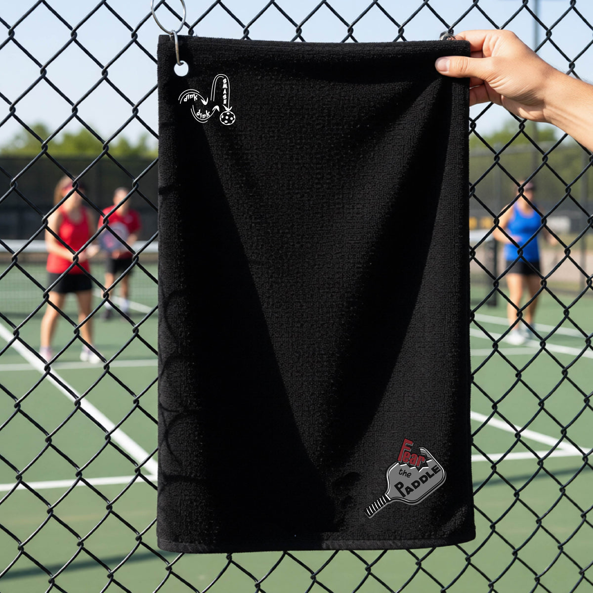 A black court towel with grommet and hook with a DINK DINK SMASH logo in white on the top left and a pickleball graphic of text 'FEAR THE PADDLE' in white cut out into a pickleball paddle on the bottom right. 