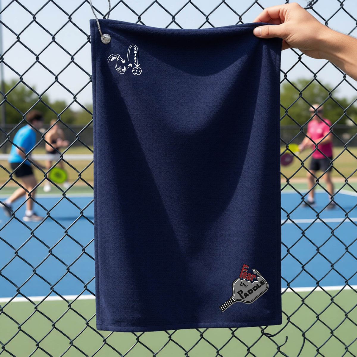 A navy blue court towel with grommet and hook with a DINK DINK SMASH logo in white on the top left and a pickleball graphic of text 'FEAR THE PADDLE' in white cut out into a pickleball paddle on the bottom right. 