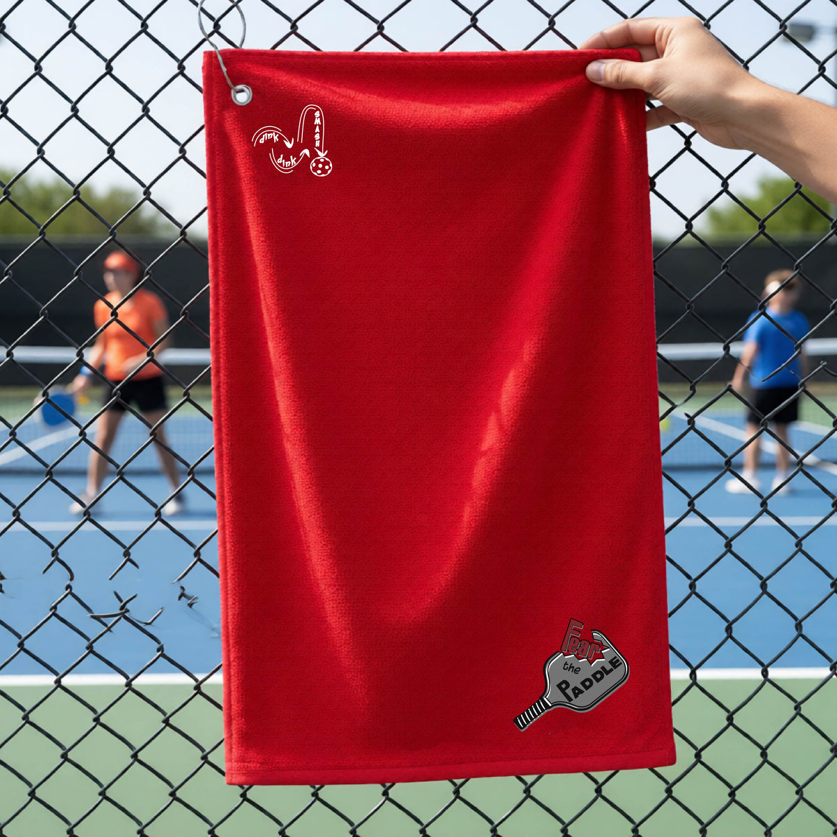 A red court towel with grommet and hook with a DINK DINK SMASH logo in white on the top left and a pickleball graphic of text 'FEAR THE PADDLE' in white cut out into a pickleball paddle on the bottom right. 