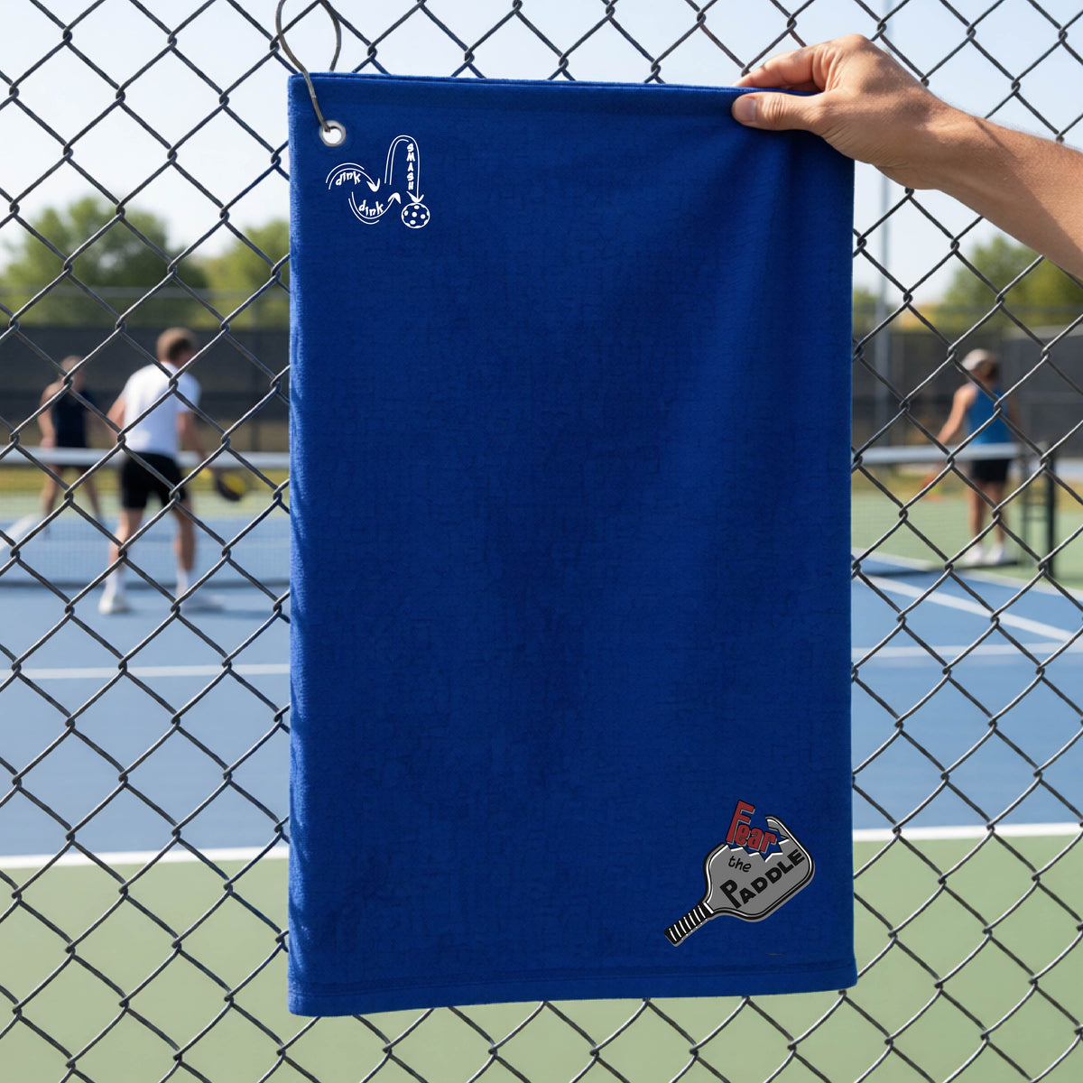 A royal blue court towel with grommet and hook with a DINK DINK SMASH logo in white on the top left and a pickleball graphic of text 'FEAR THE PADDLE' in white cut out into a pickleball paddle on the bottom right. 