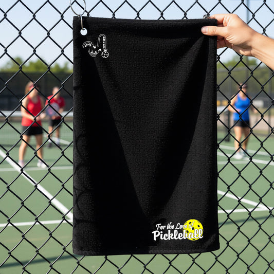 A close-up of a black court towel with grommet and hook and a pickleball graphic of ‘FOR THE LOVE OF PICKLEBALL’ in white with a yellow pickleball behind it, and a DINK DINK SMASH logo in white on the top of the towel.