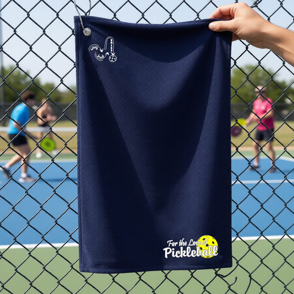 A close-up of a navy blue court towel with grommet and hook and a pickleball graphic of ‘FOR THE LOVE OF PICKLEBALL’ in white with a yellow pickleball behind it, and a DINK DINK SMASH logo in white on the top of the towel.