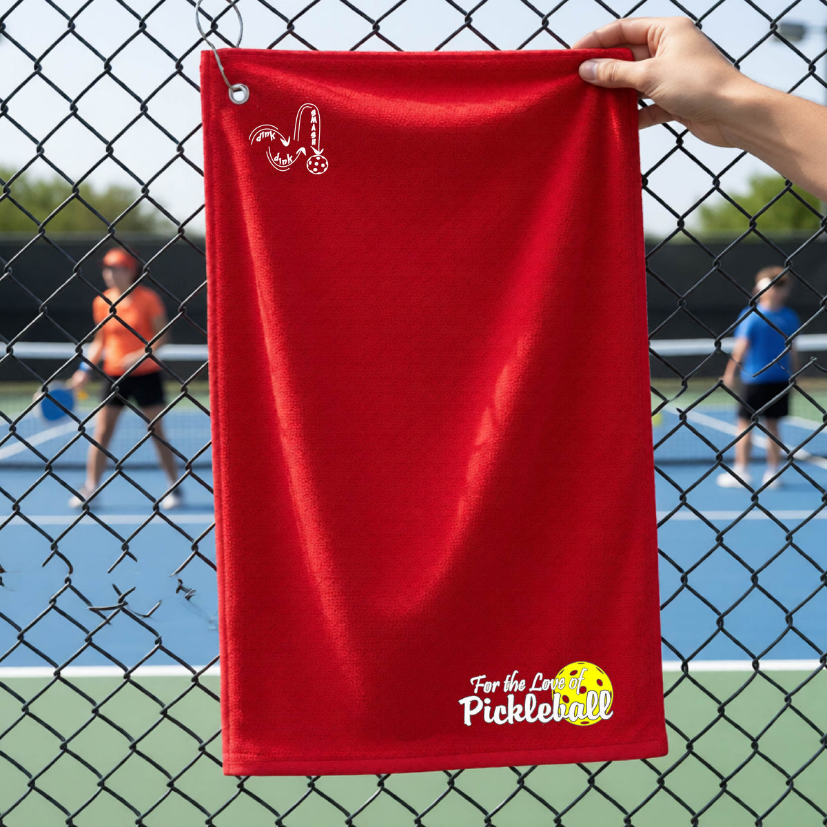 A close-up of a red court towel with grommet and hook and a pickleball graphic of ‘FOR THE LOVE OF PICKLEBALL’ in white with a yellow pickleball behind it, and a DINK DINK SMASH logo in white on the top of the towel.