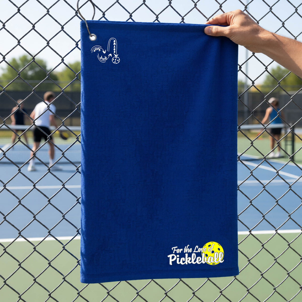A close-up of a royal blue court towel with grommet and hook and a pickleball graphic of ‘FOR THE LOVE OF PICKLEBALL’ in white with a yellow pickleball behind it, and a DINK DINK SMASH logo in white on the top of the towel.