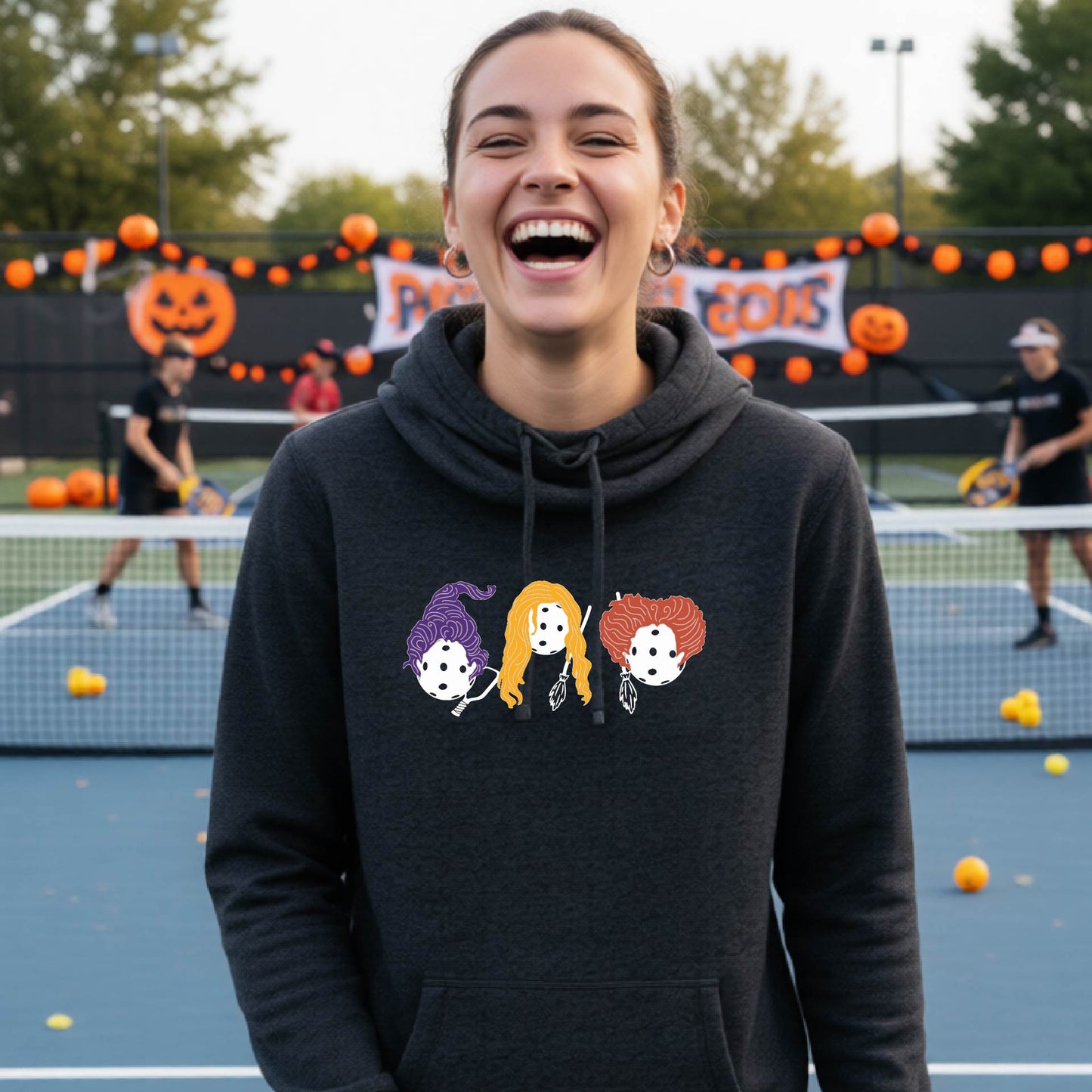 A woman smiling on a tennis court wearing a dark charcoal gray hoodie with a graphic of three pickleball halloween Hocus Pocus cartoon characters wearing purple, yellow, and orange hair with dice heads.