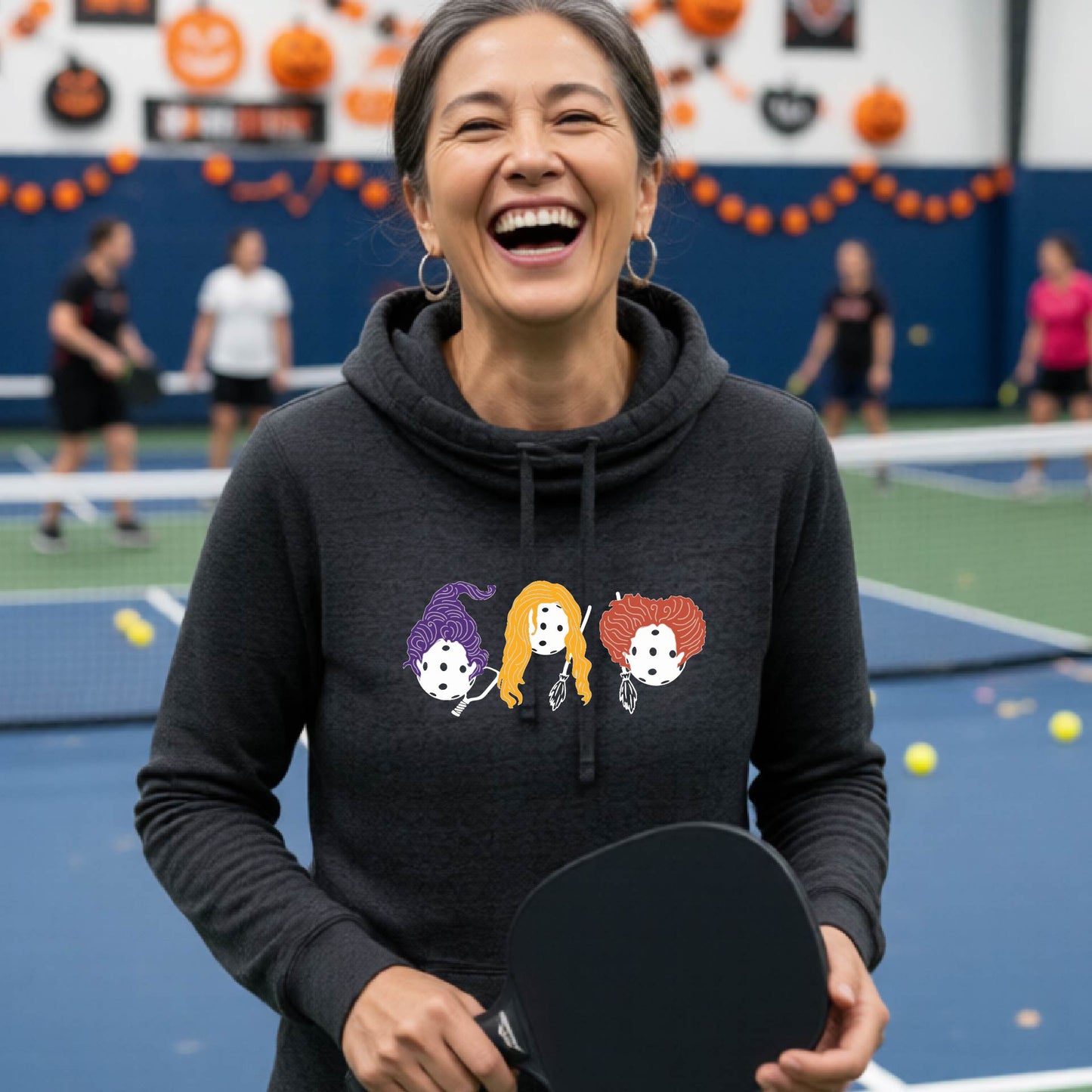 A woman laughing on an indoor pickleball court wearing a dark gray hoodie with a colorful graphic of three cartoon pickleball halloween Hocus pocus , holding a paddle.