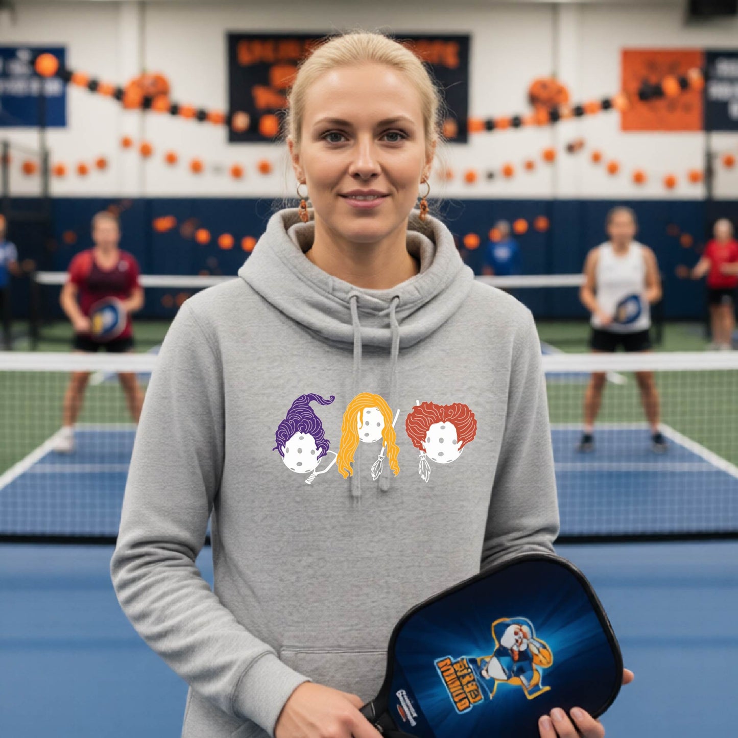 A woman stands on an indoor pickleball court wearing a heather gray hoodie with a colorful pickleball hocus Pocus cartoon graphic on the chest and holding a blue pickleball paddle. Other players and court decorations are visible in the background.