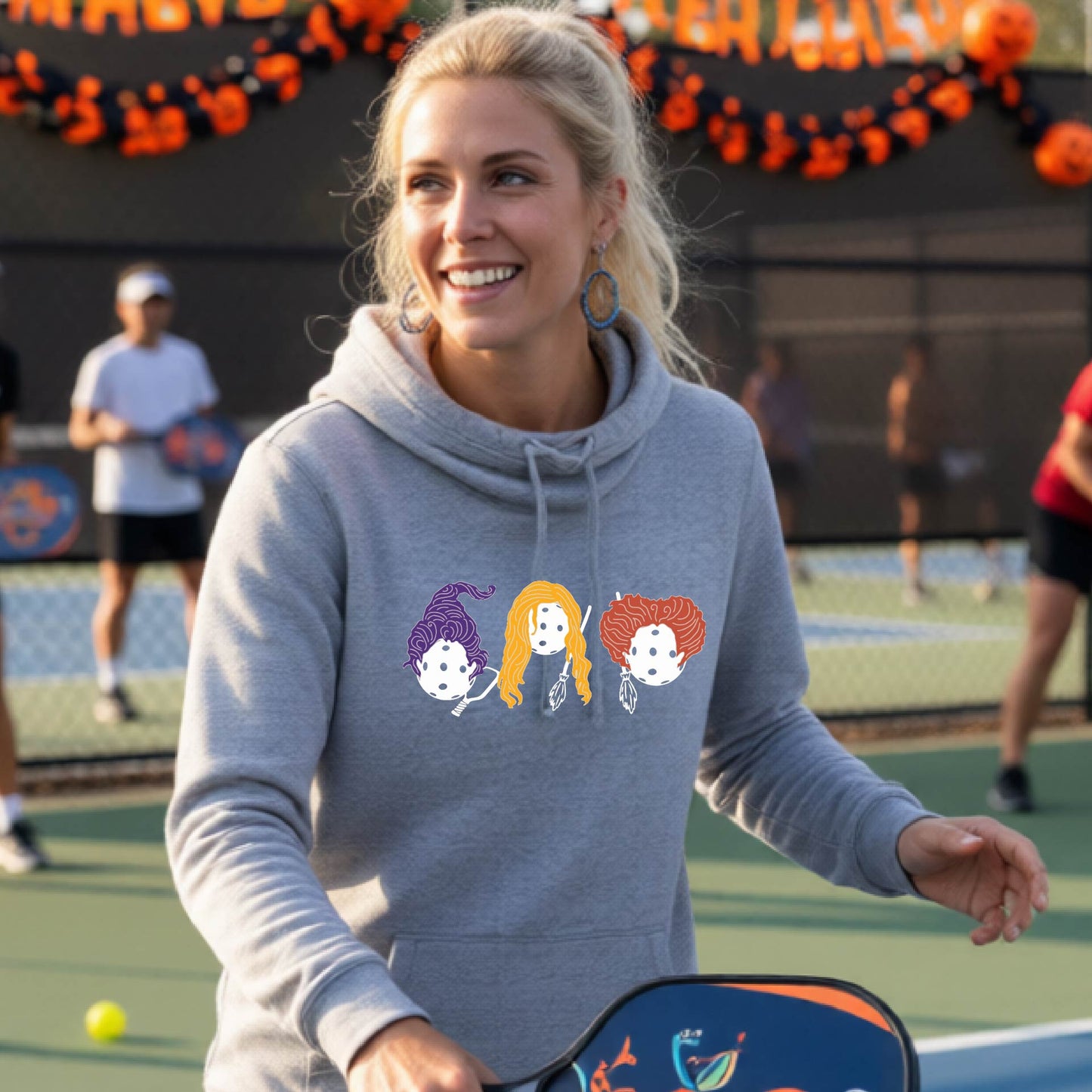 A woman on an outdoor pickleball court wearing a heather gray hoodie with a graphic of three cartoon pickleball halloween Hocus pocus girls holding pickleballs. The background shows a pickleball court with other players and orange Halloween decorations.