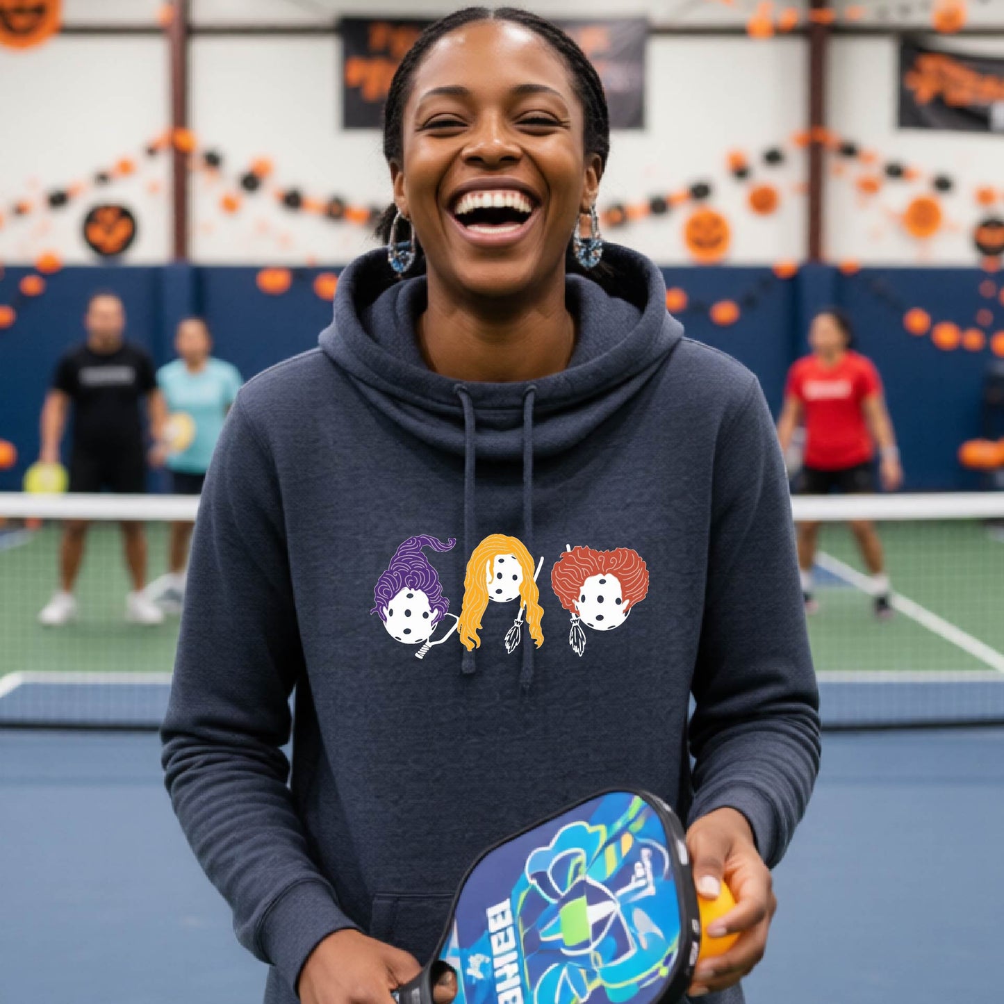 A woman smiling in a gym while wearing a navy blue hoodie with a graphic of three cartoon pickleball halloween Hocus holding a pickleball paddle.