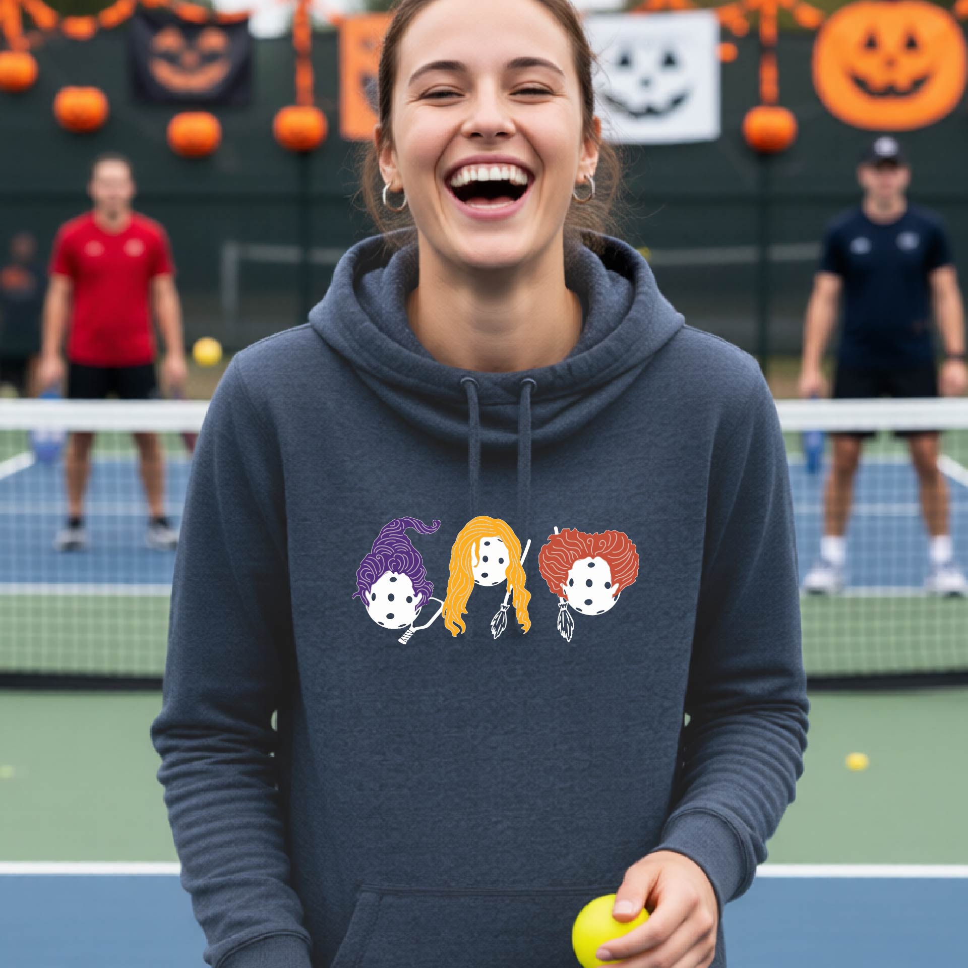 A woman on a tennis court wearing a navy blue hoodie with a graphic of three cartoon pickleball halloween Hocus Pocus witch characters with purple, orange, and red hair. The background shows other players on the court and Halloween decorations.