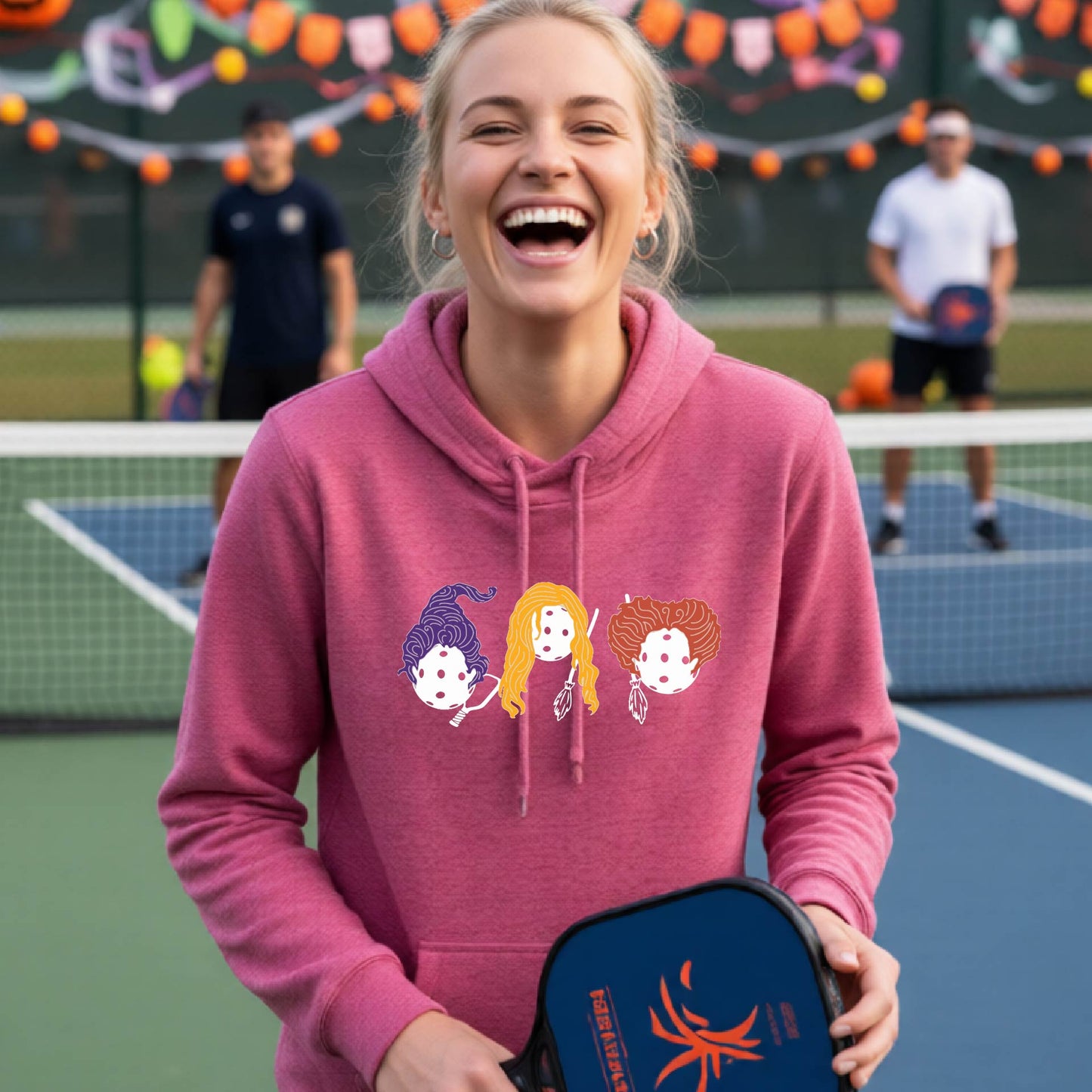 A woman laughing on a pickleball court wearing a pink hoodie with a graphic of three cartoon pickleball halloween Hocus holding a blue pickleball paddle. The background shows other players and a decorated court.