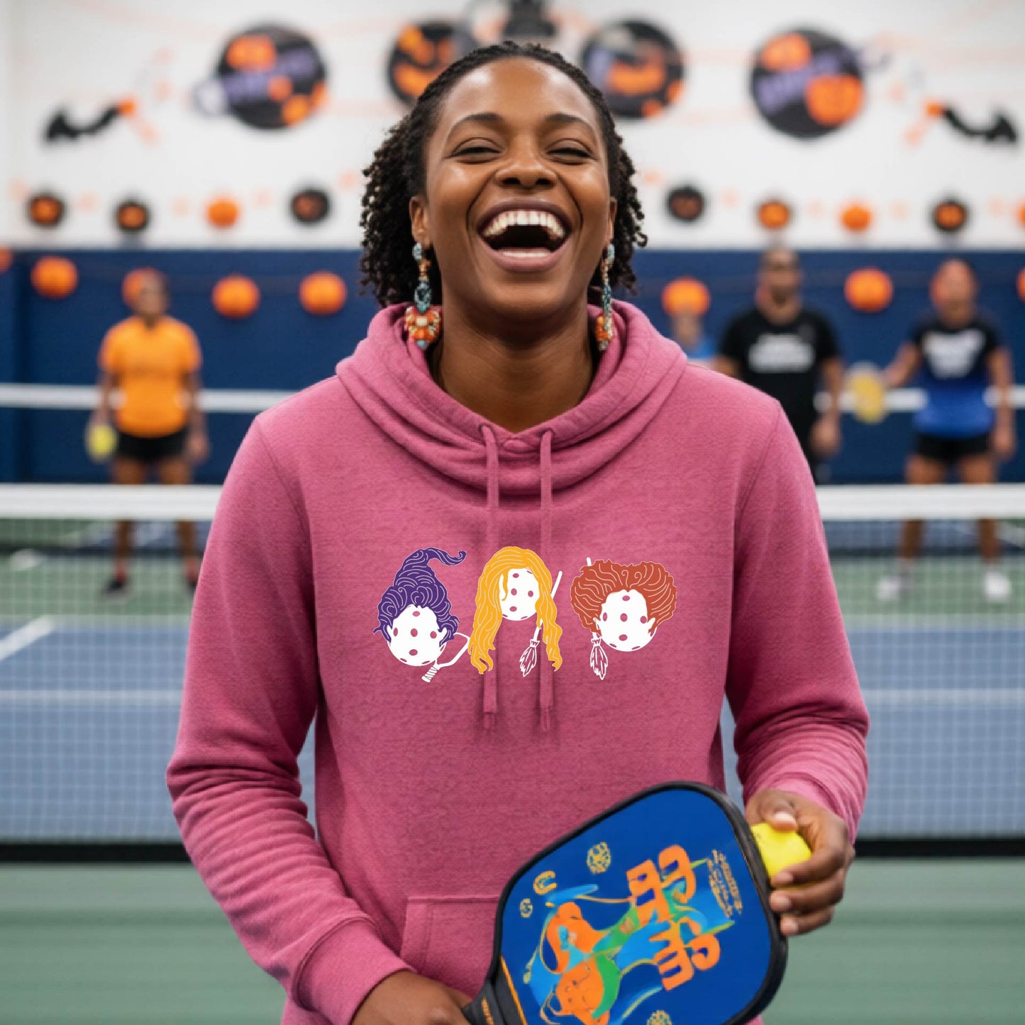 A woman laughing in a pickleball court wearing a pickleball halloween Hocus Pocus design.  She is holding a blue pickleball paddle and a yellow ball, with other players and a court visible in the background.