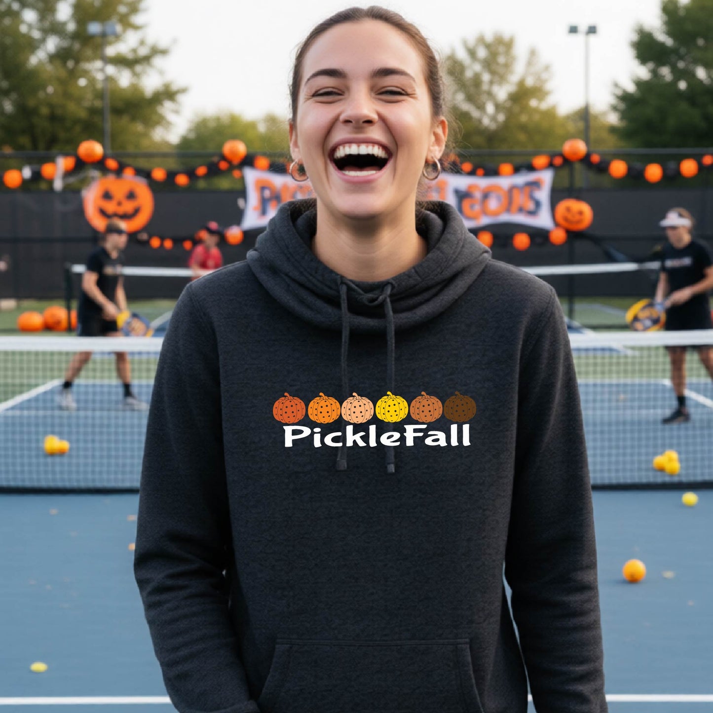 A woman stands on a ickleball court wearing a heather black cowl-neck hoodie with a 'PickleFall' graphic and six pickelball pumpkins on the design with court Halloween decorations visible in the background.
