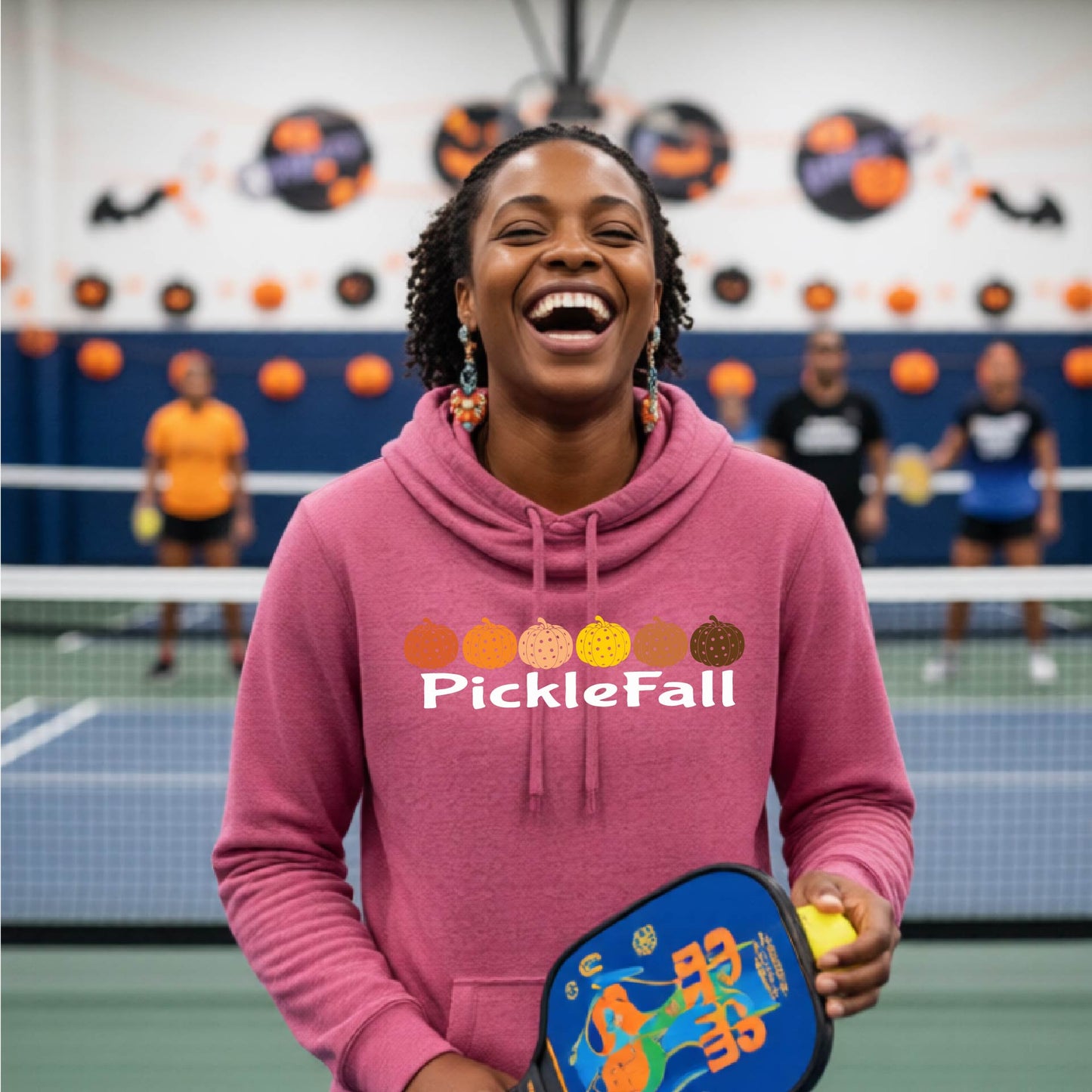 A woman stands on a ickleball court wearing a pink cowl-neck hoodie with a 'PickleFall' graphic and six pickelball pumpkins on the design with court Halloween decorations visible in the background.