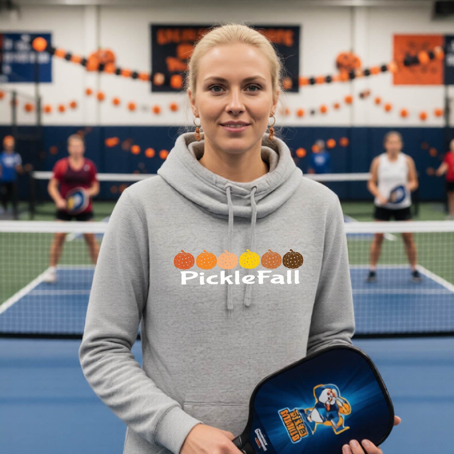 A woman stands on a ickleball court wearing a gray cowl-neck hoodie with a 'PickleFall' graphic and six pickelball pumpkins on the design with court Halloween decorations visible in the background.