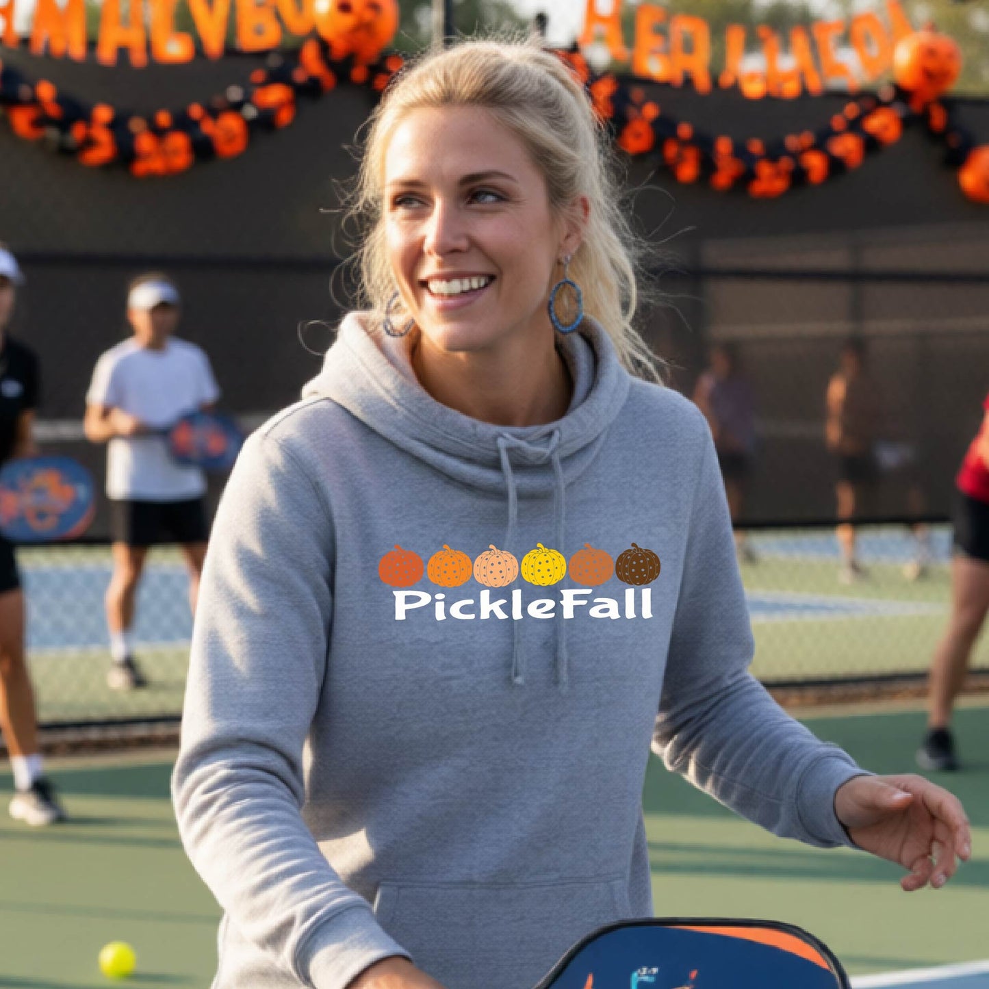 A woman stands on a ickleball court wearing a gray cowl-neck hoodie with a 'PickleFall' graphic and six pickelball pumpkins on the design with court Halloween decorations visible in the background.