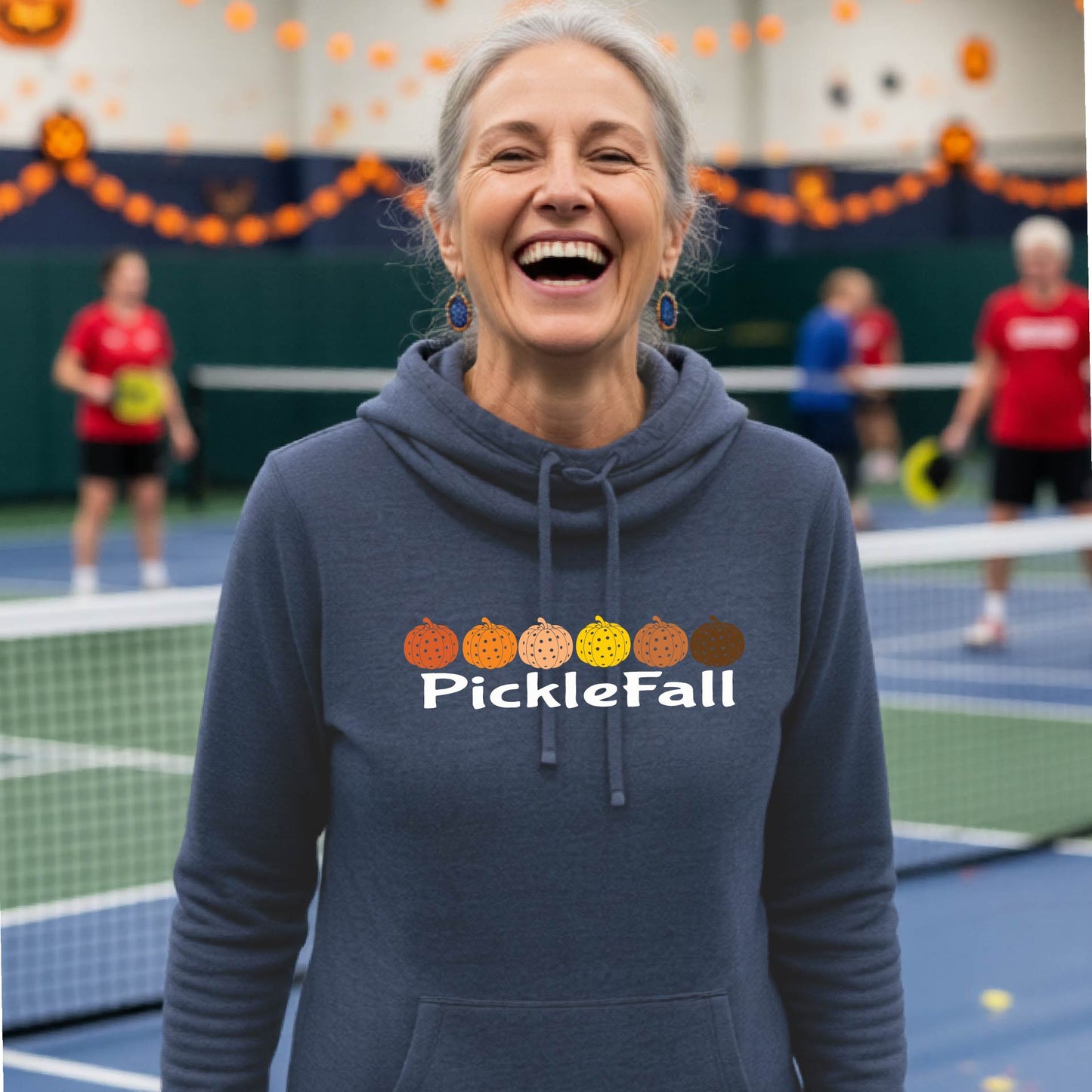 A woman stands on a ickleball court wearing a heather navy blue cowl-neck hoodie with a 'PickleFall' graphic and six pickelball pumpkins on the design with court Halloween decorations visible in the background.