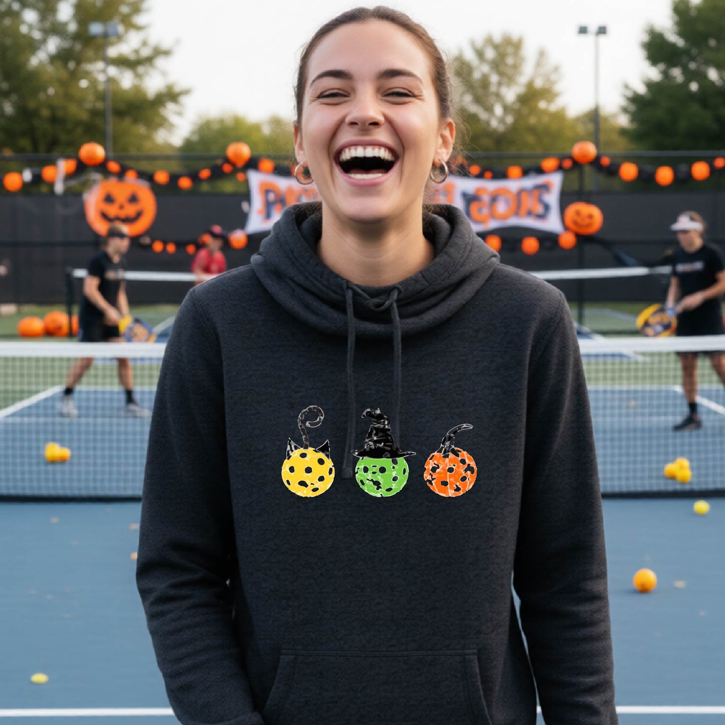 A woman on a pickleball court wearing a black heathered cowl-neck hoodie with three colorful pickleball pumpkin graphics on the chest. The background shows other players and orange Halloween decorations.