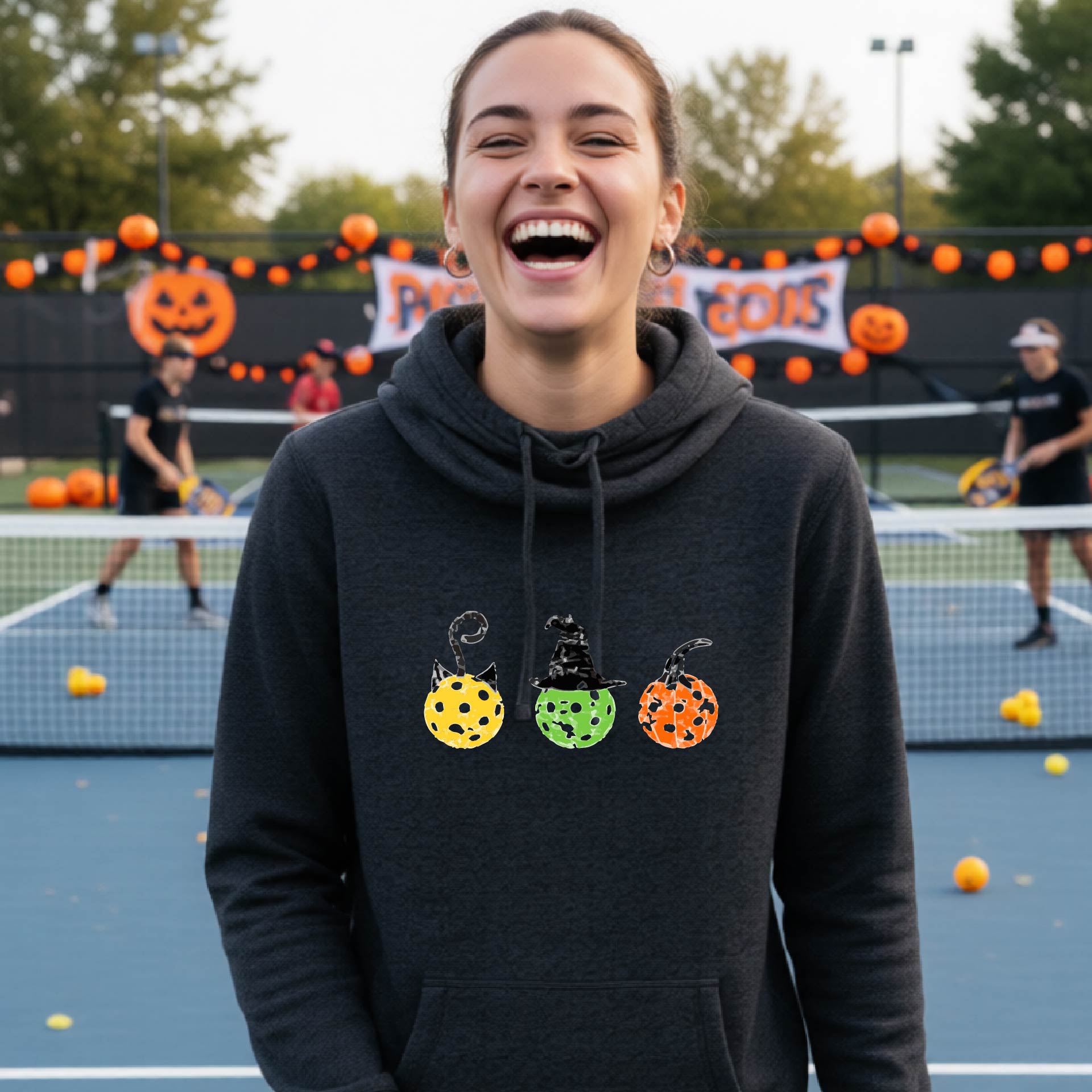 A woman on a pickleball court wearing a black heathered cowl-neck hoodie with three colorful pickleball pumpkin graphics on the chest. The background shows other players and orange Halloween decorations.