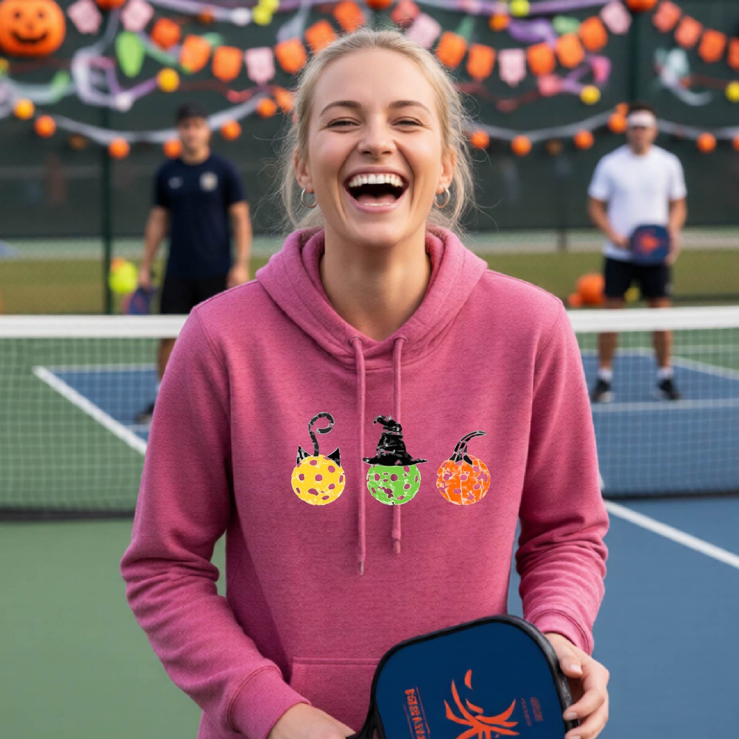 A woman on a pickleball court wearing a pink cowl-neck hoodie with three colorful pickleball pumpkin graphics on the chest. The background shows other players and orange Halloween decorations.background.