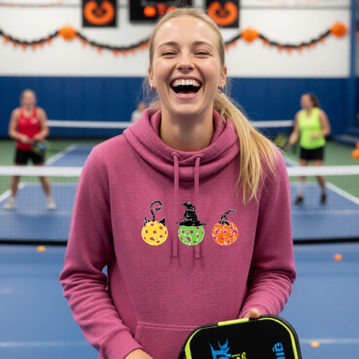A woman on a pickleball court wearing a pink cowl-neck hoodie with three colorful pickleball pumpkin graphics on the chest. The background shows other players and orange Halloween decorations.