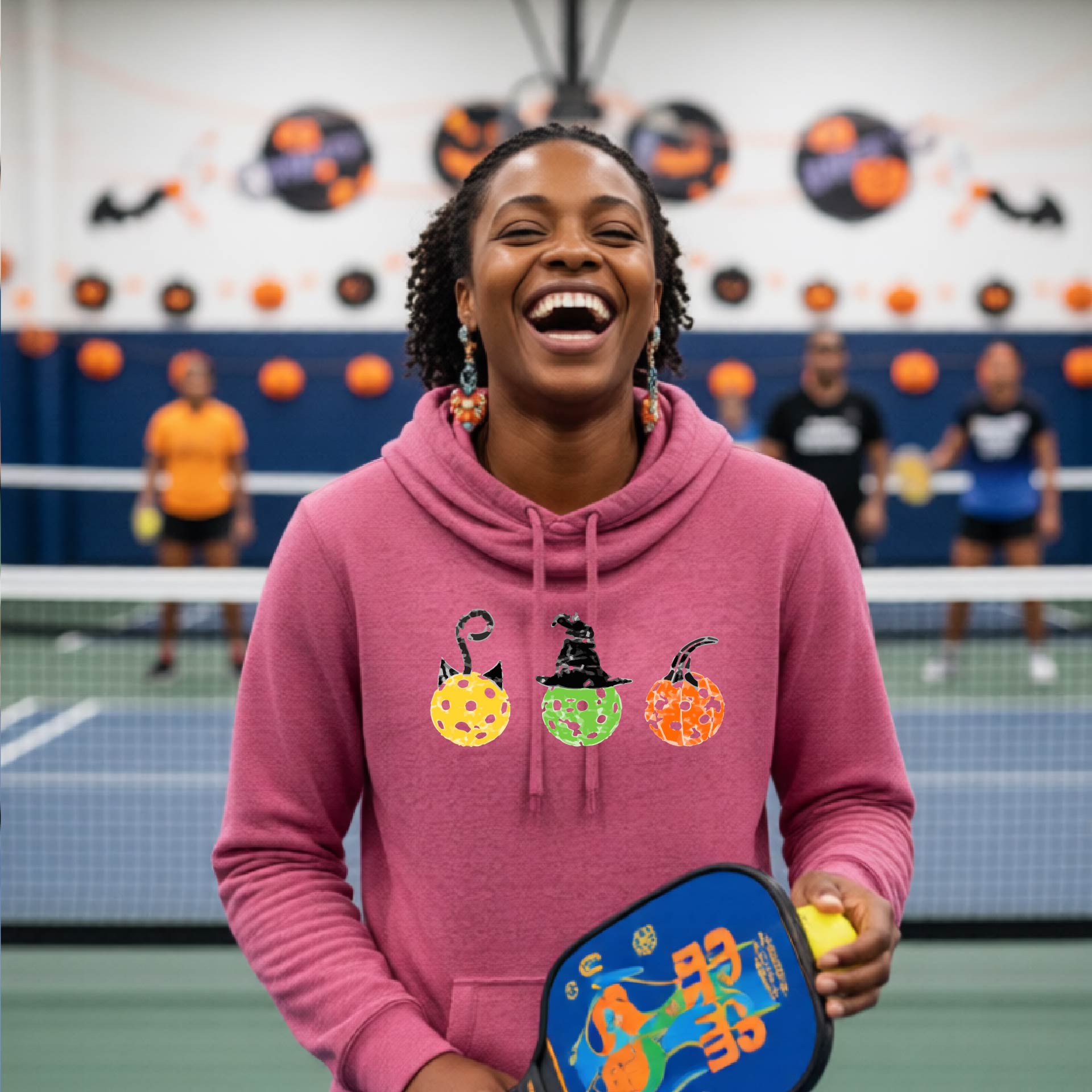 A woman on a pickleball court wearing a pink cowl-neck hoodie with three colorful pickleball pumpkin graphics on the chest. The background shows other players and orange Halloween decorations.