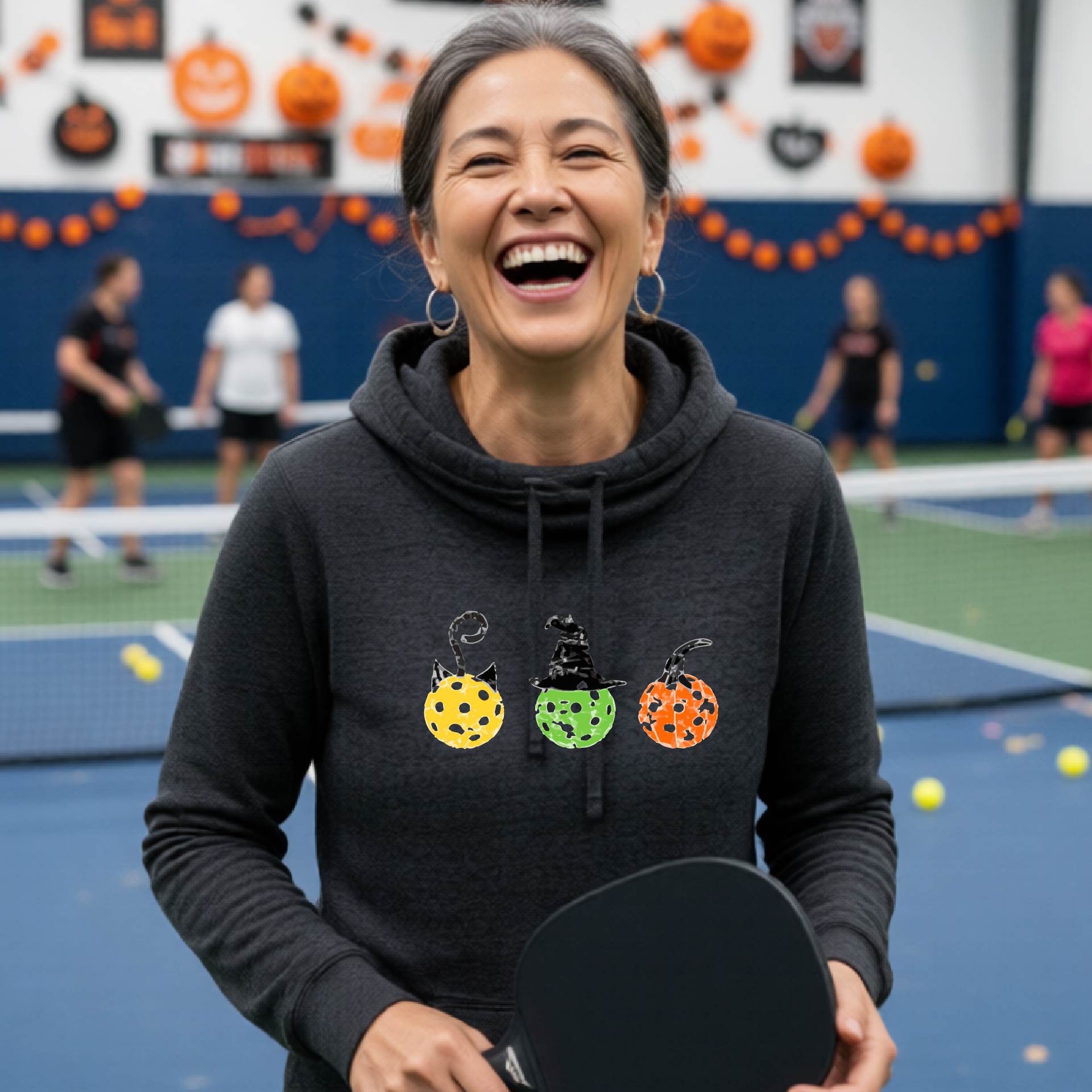 A woman on a pickleball court wearing a black heatherd cowl-neck hoodie with three colorful pickleball pumpkin graphics on the chest. The background shows other players and orange Halloween decorations.decorations.