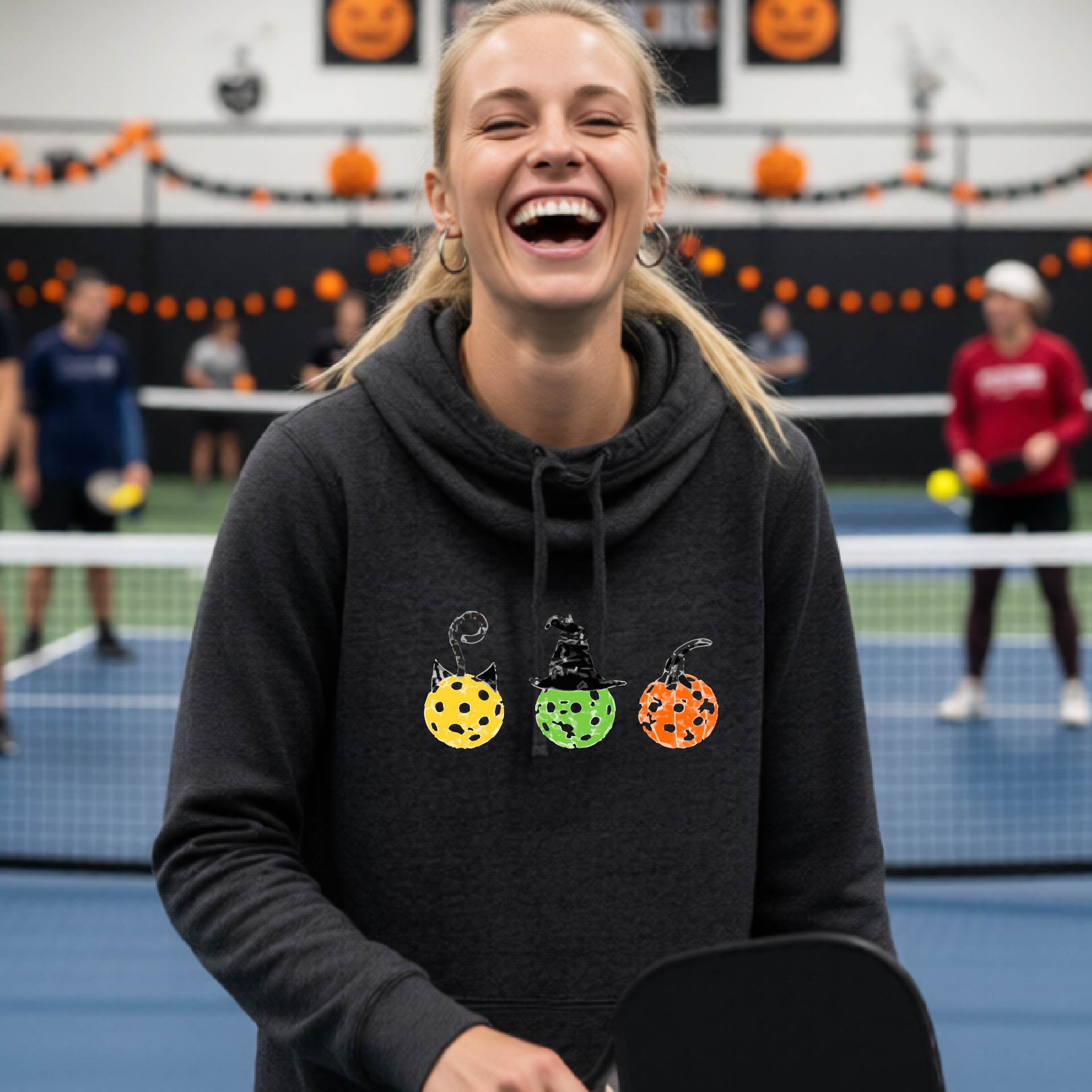 A woman on a pickleball court wearing a black cowl-neck hoodie with three colorful pickleball pumpkin graphics on the chest. The background shows other players and orange Halloween decorations.