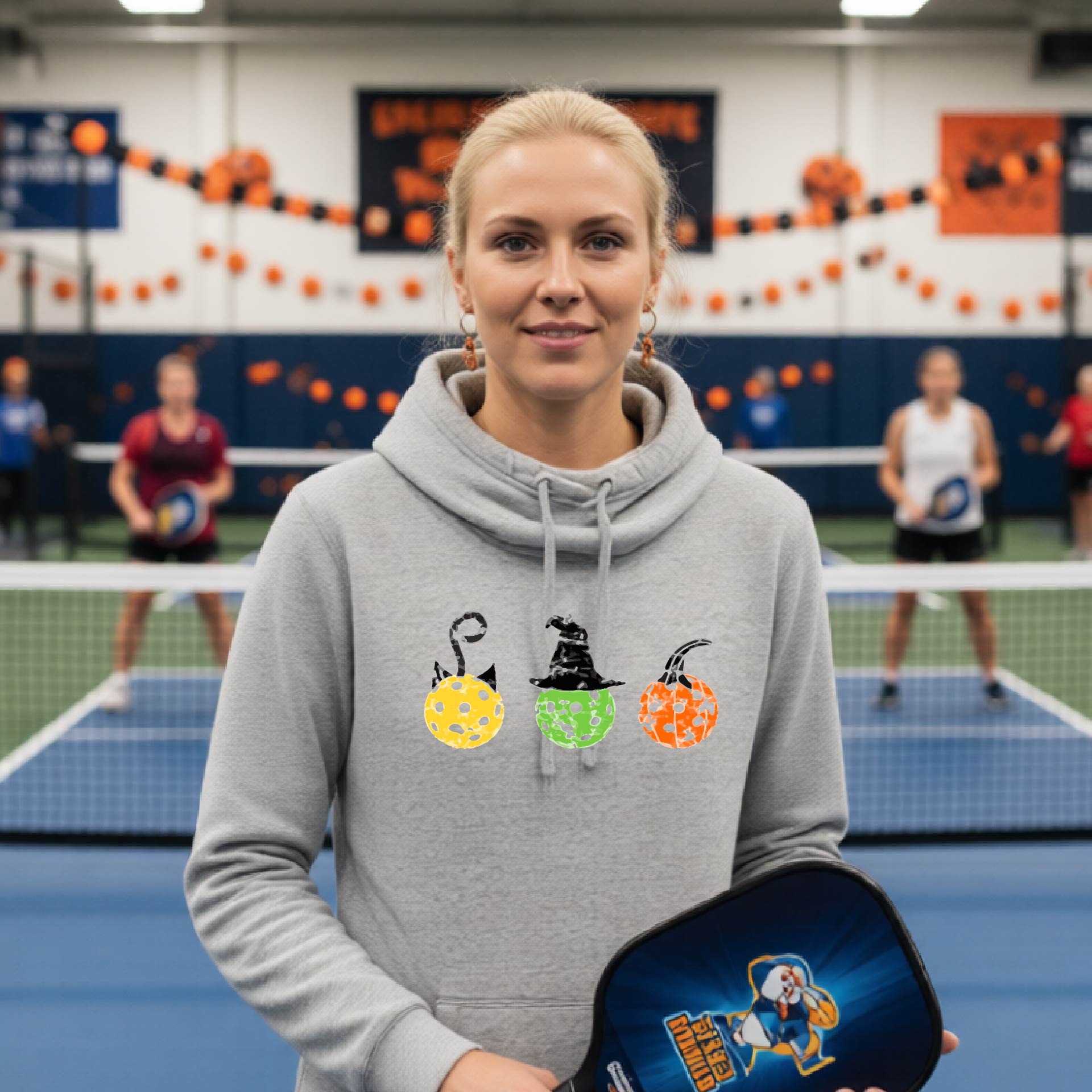 A woman on a pickleball court wearing a light gray cowl-neck hoodie with three colorful pickleball pumpkin graphics on the chest. The background shows other players and orange Halloween decorations.