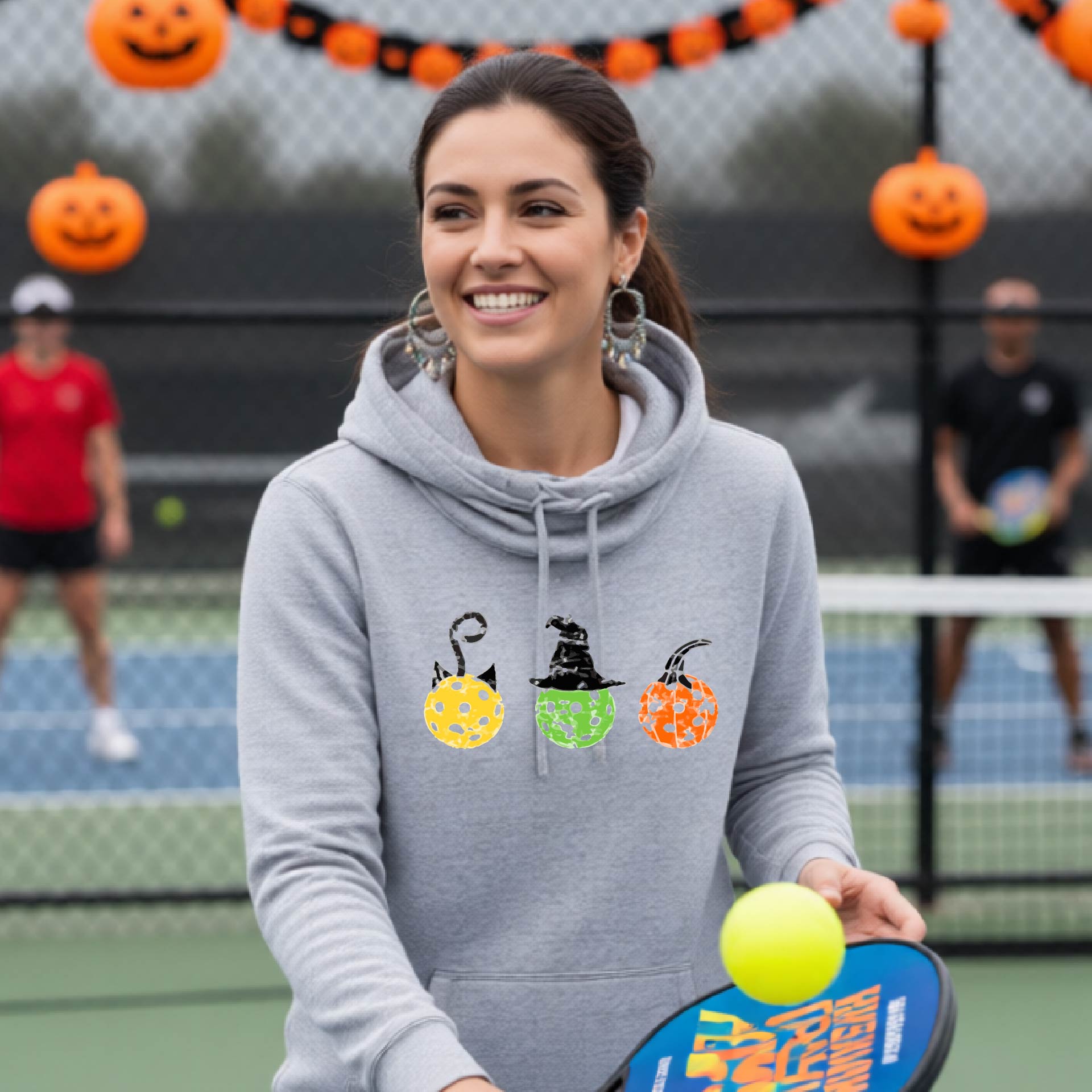 A woman on a pickleball court wearing a light grey cowl-neck hoodie with three colorful pickleball pumpkin graphics on the chest. The background shows other players and orange Halloween decorations.