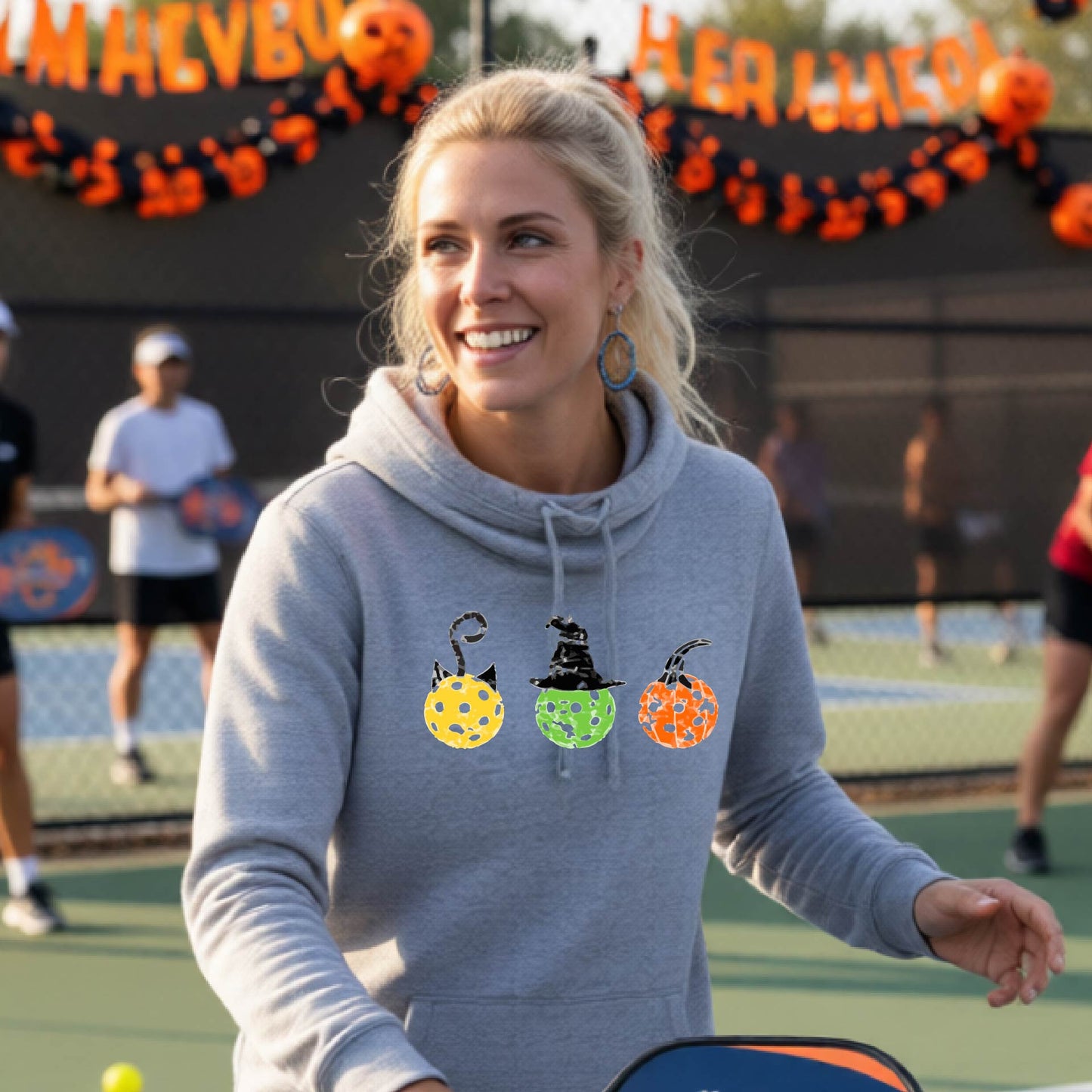 A woman on a pickleball court wearing a heather gray hoodie with three colorful pickleball pumpkin graphics on the chest. The background shows other players and orange Halloween decorations.