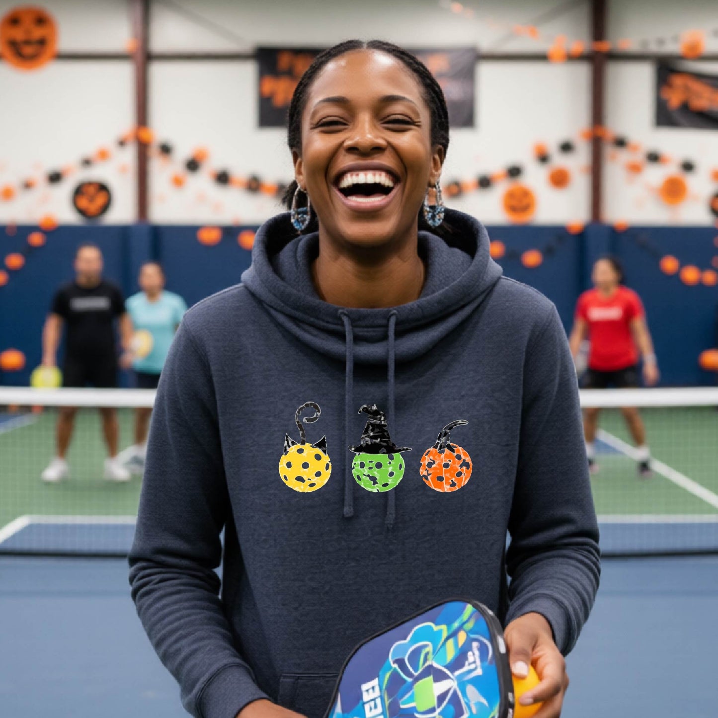 A woman on a pickleball court wearing a navy blue cowl-neck hoodie with three colorful pickleball pumpkin graphics on the chest. The background shows other players and orange Halloween decorations.