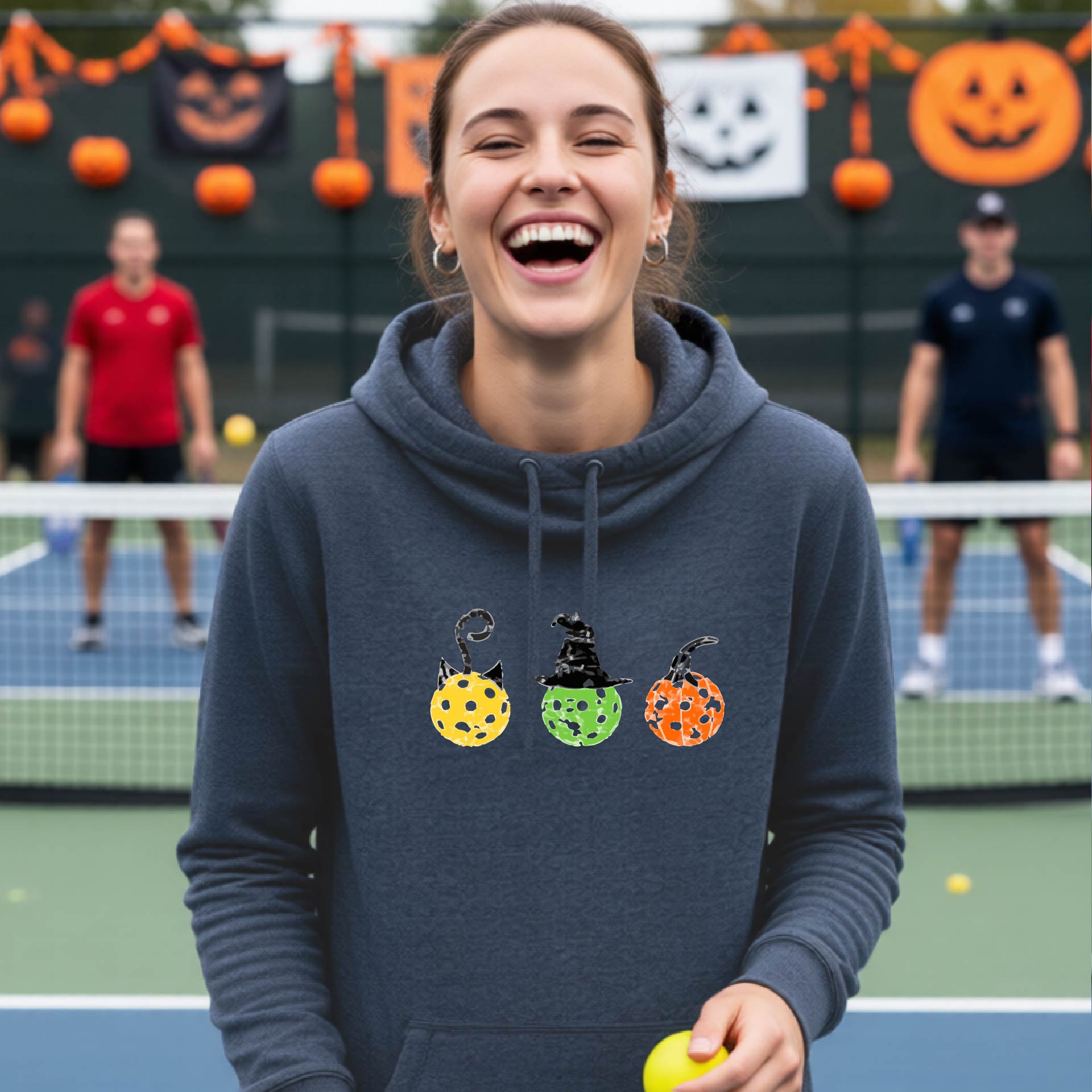 A woman on a pickleball court wearing a navy blue cowl-neck hoodie with three colorful pickleball pumpkin graphics on the chest. The background shows other players and orange Halloween decorations.