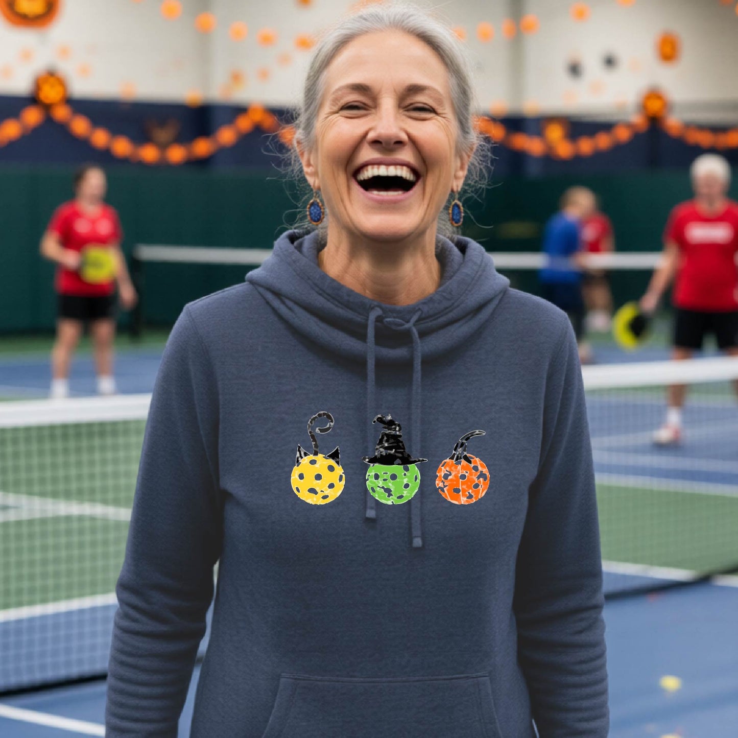 A woman on a pickleball court wearing a navy blue cowl-neck hoodie with three colorful pickleball pumpkin graphics on the chest. The background shows other players and orange Halloween decorations.background.