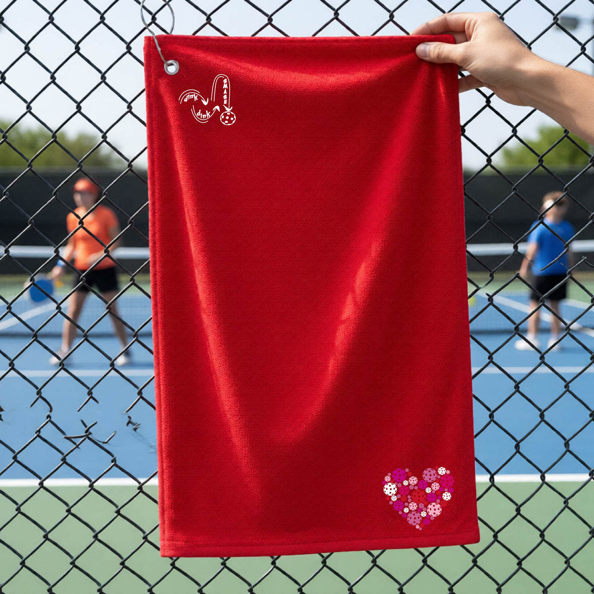 A red textured terry cloth towel is held up by a hand in front of a chain-link fence with a pickleball court in the background. The towel features a logo of 'DINK DINK SMASH'  near the top left and a heart-shaped  patch made of pickleballs near the bottom right.