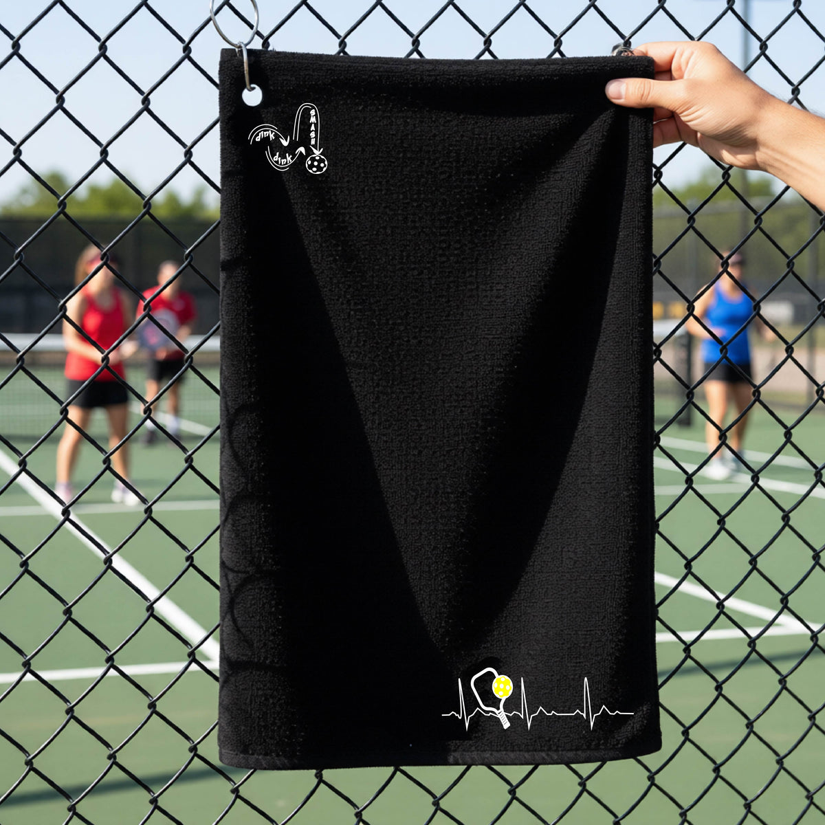 A close-up of a black grommet and hook terry cloth court towels porting a white pickleball graphic of a heartbeat EKG including a pickleball paddle and a yellow pickleball across the check in the botton right dorner and a 'DINK DINK SMASH' logo in the upper left corner, in front of a pickleball court.
