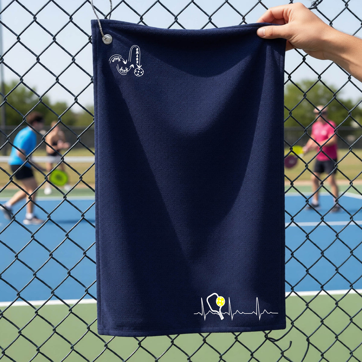 A close-up of a navy blue grommet and hook terry cloth court towels porting a white pickleball graphic of a heartbeat EKG including a pickleball paddle and a yellow pickleball across the check in the botton right dorner and a 'DINK DINK SMASH' logo in the upper left corner, in front of a pickleball court.