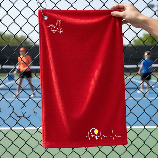 A close-up of a red grommet and hook terry cloth court towels porting a white pickleball graphic of a heartbeat EKG including a pickleball paddle and a yellow pickleball across the check in the botton right dorner and a 'DINK DINK SMASH' logo in the upper left corner, in front of a pickleball court.