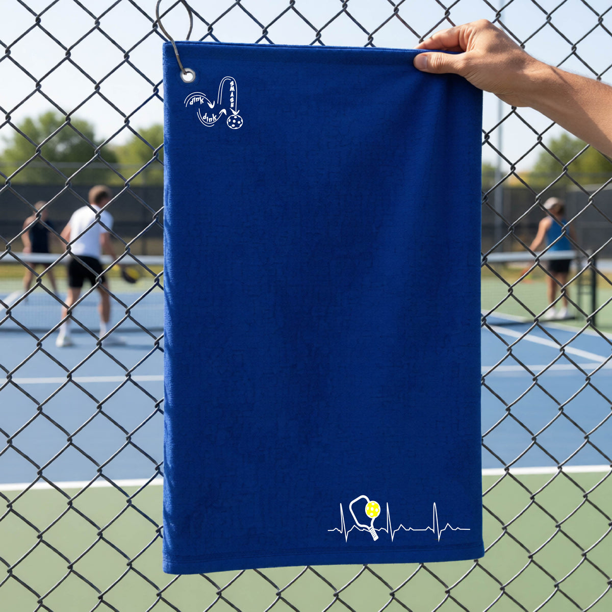 A close-up of a royal blue grommet and hook terry cloth court towels porting a white pickleball graphic of a heartbeat EKG including a pickleball paddle and a yellow pickleball across the check in the botton right dorner and a 'DINK DINK SMASH' logo in the upper left corner, in front of a pickleball court.