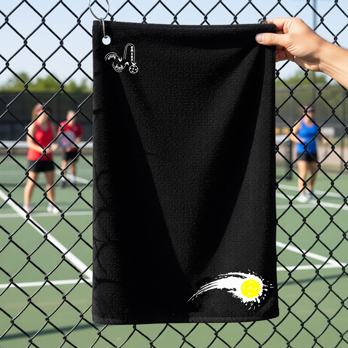 A close-up of a black terry cloth court towel on a pickleballc ourt fence with a white pickleball graphic of a yellow pickleball with a motion trail effect, displayed in the lower right corner and a white logo of DINK DINK SMASH in the upper left corner.  
