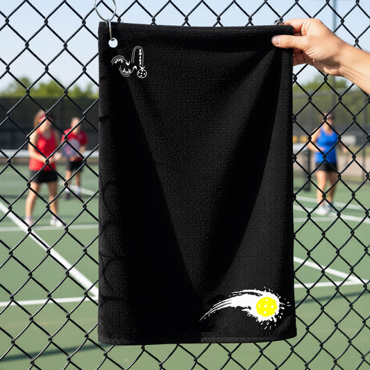 A close-up of a black terry cloth court towel on a pickleballc ourt fence with a white pickleball graphic of a yellow pickleball with a motion trail effect, displayed in the lower right corner and a white logo of DINK DINK SMASH in the upper left corner.  