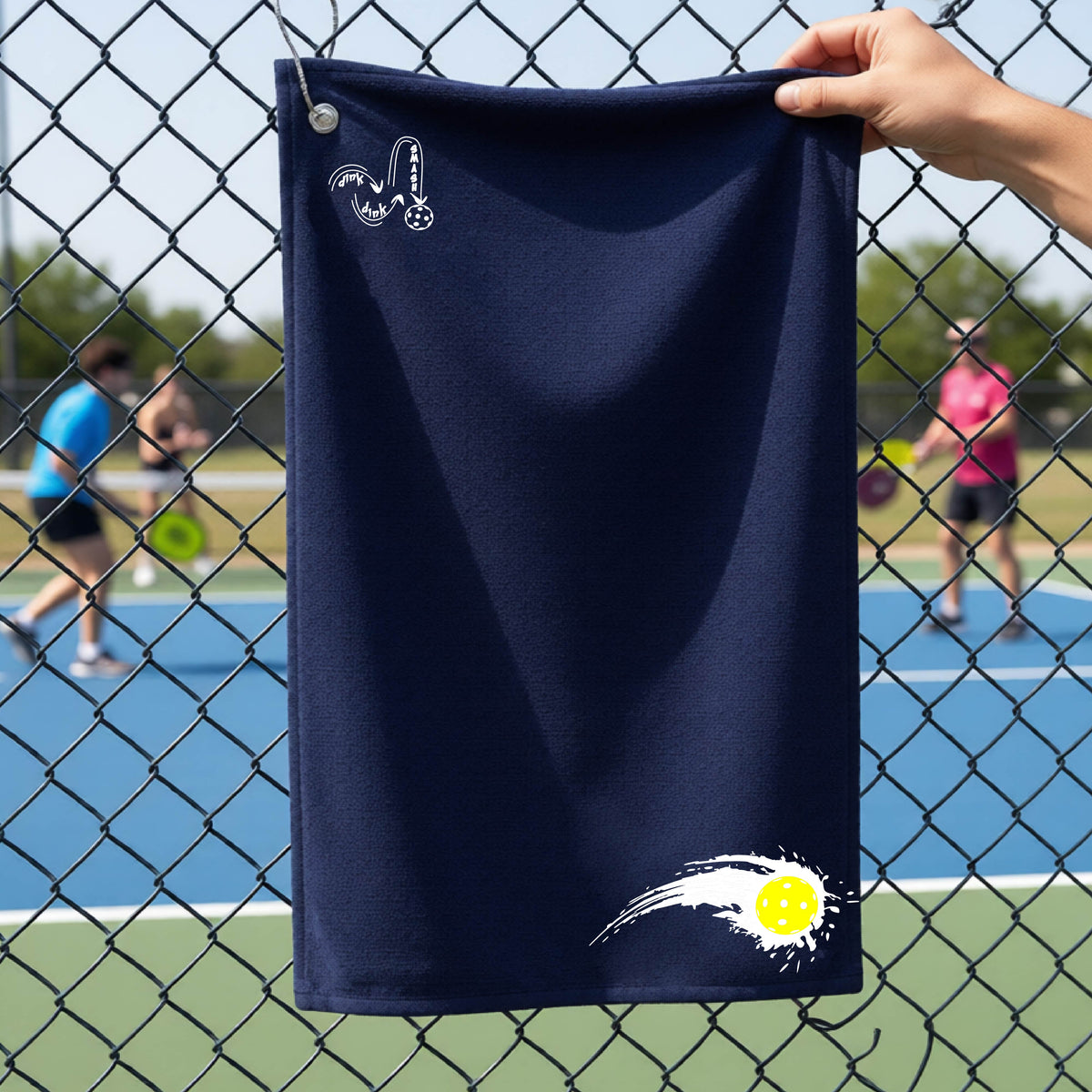 A close-up of a navy blue terry cloth court towel on a pickleballc ourt fence with a white pickleball graphic of a yellow pickleball with a motion trail effect, displayed in the lower right corner and a white logo of DINK DINK SMASH in the upper left corner.  