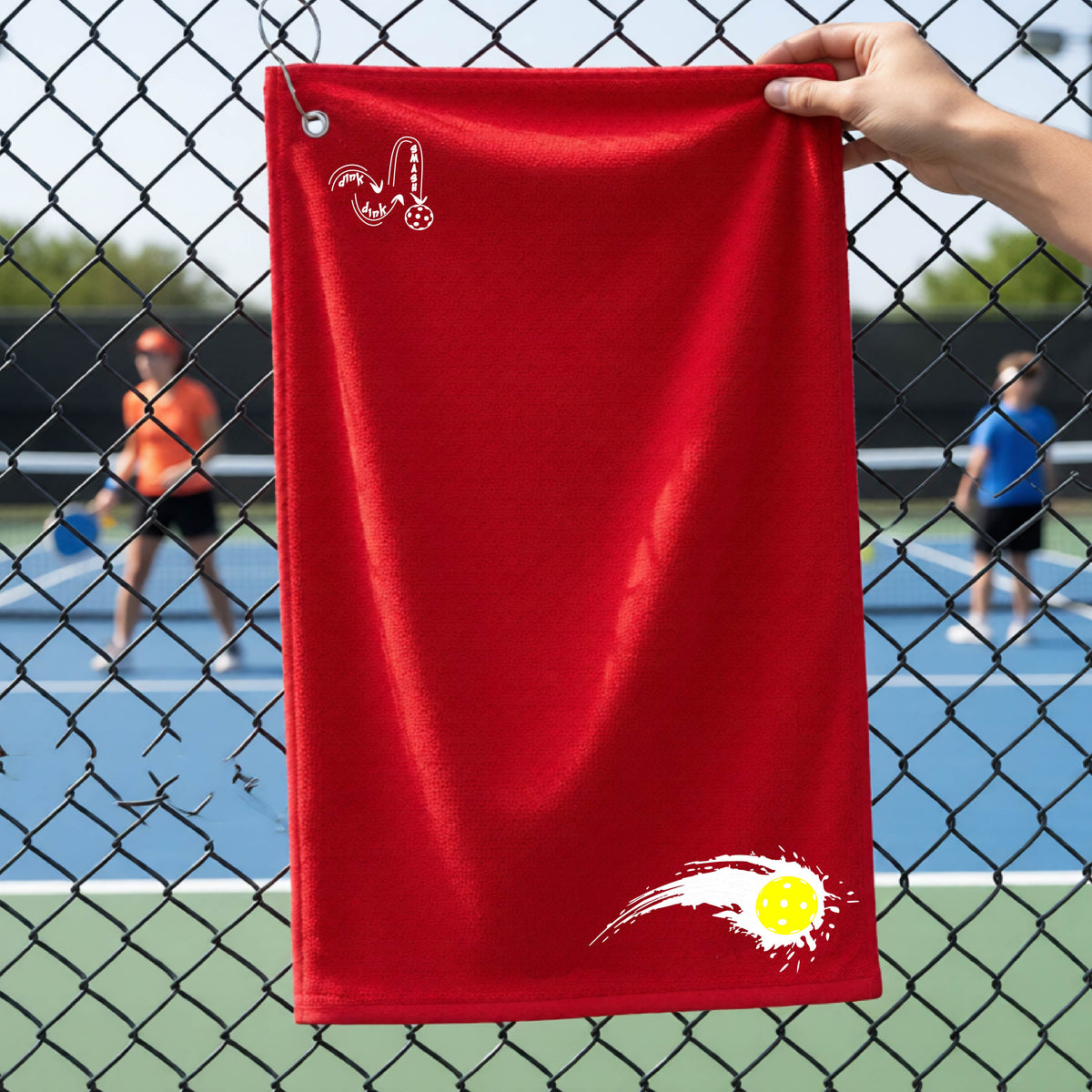 A close-up of a red terry cloth court towel on a pickleballc ourt fence with a white pickleball graphic of a yellow pickleball with a motion trail effect, displayed in the lower right corner and a white logo of DINK DINK SMASH in the upper left corner.  