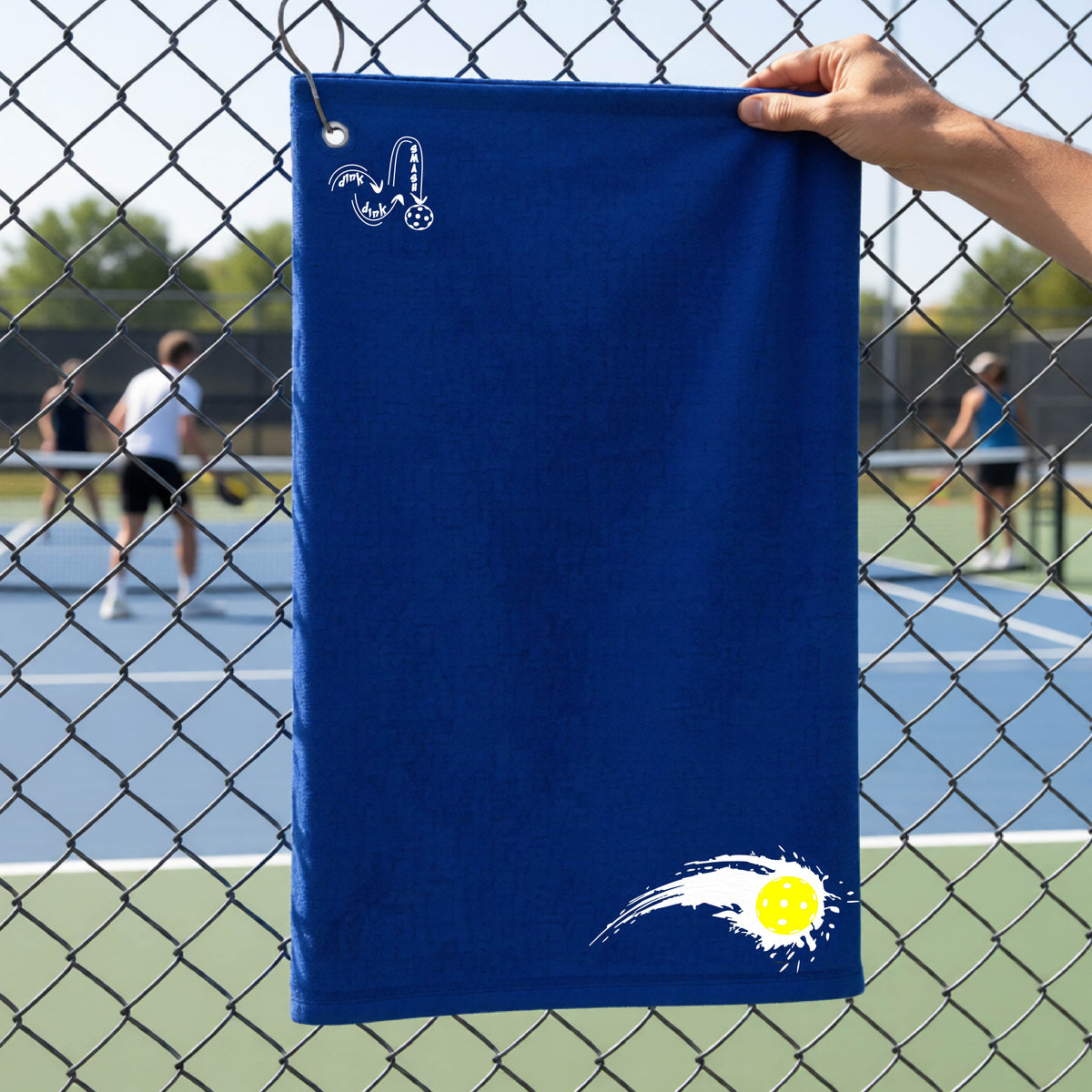 A close-up of a royal blue terry cloth court towel on a pickleballc ourt fence with a white pickleball graphic of a yellow pickleball with a motion trail effect, displayed in the lower right corner and a white logo of DINK DINK SMASH in the upper left corner.  