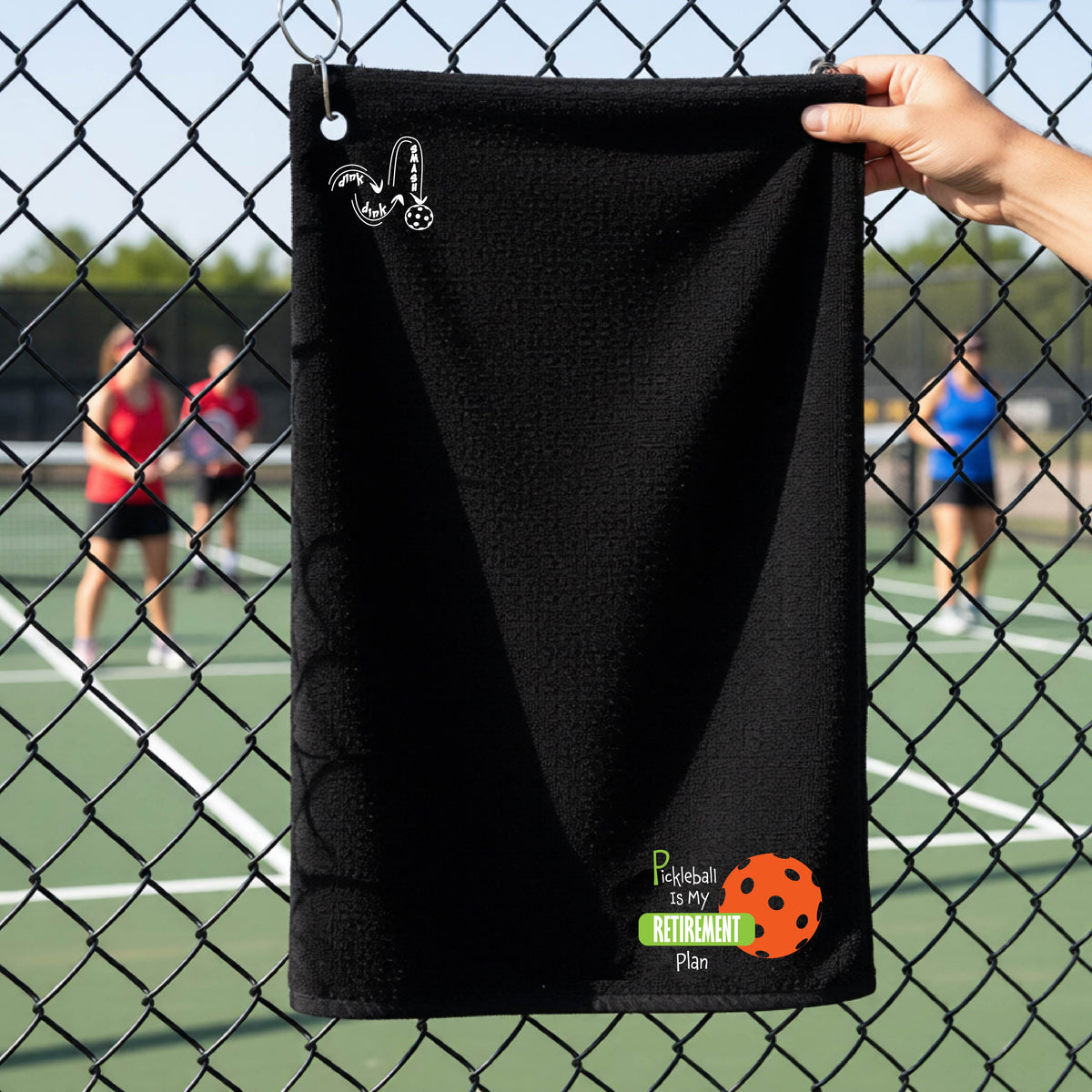 A close-up of a black terry cloth grommet court towel on a pickleball court, with a graphic of the words “Pickleball is my Retirement Plan” in green and white with an orange pickleball illustration in the background.