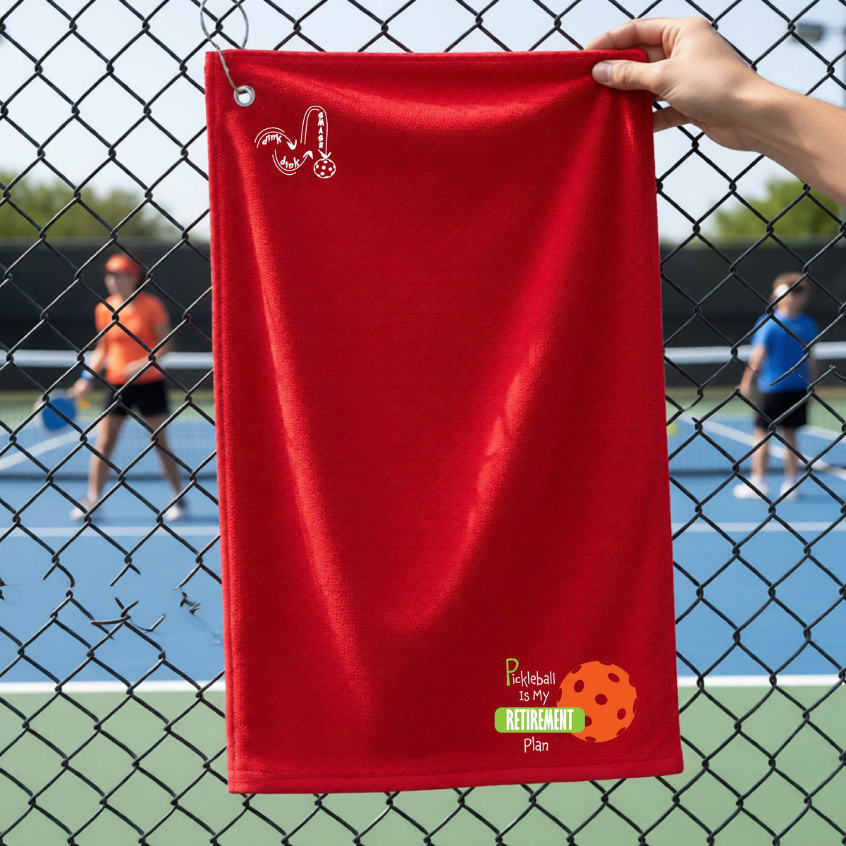 A close-up of a red terry cloth grommet court towel on a pickleball court, with a graphic of the words “Pickleball is my Retirement Plan” in green and white with an orange pickleball illustration in the background.